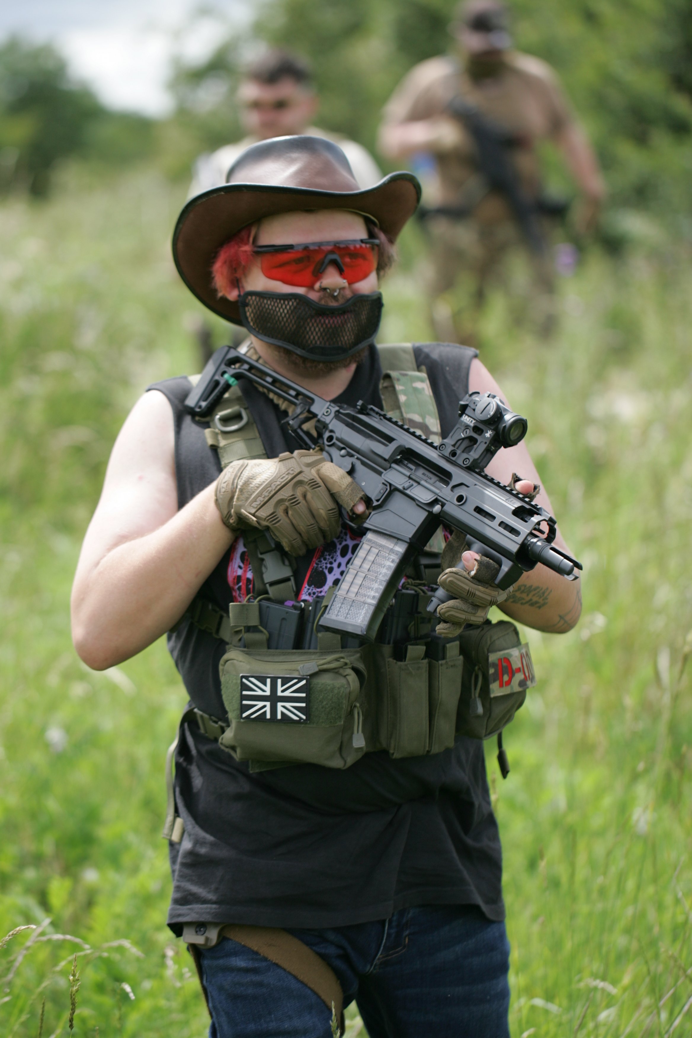 A person with red hair, wearing a cowboy hat, red sunglasses, a face mask, and tactical gear, holding a rifle outdoors in a grassy, wooded area. Two other individuals are visible blurred in the background.