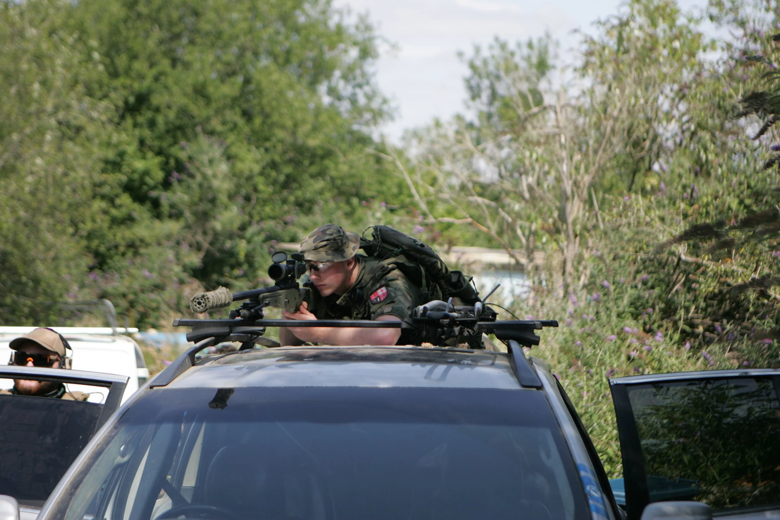 A soldier in camouflage uniform aiming a sniper rifle while on top of a black vehicle, with trees and purple flowers in the background.