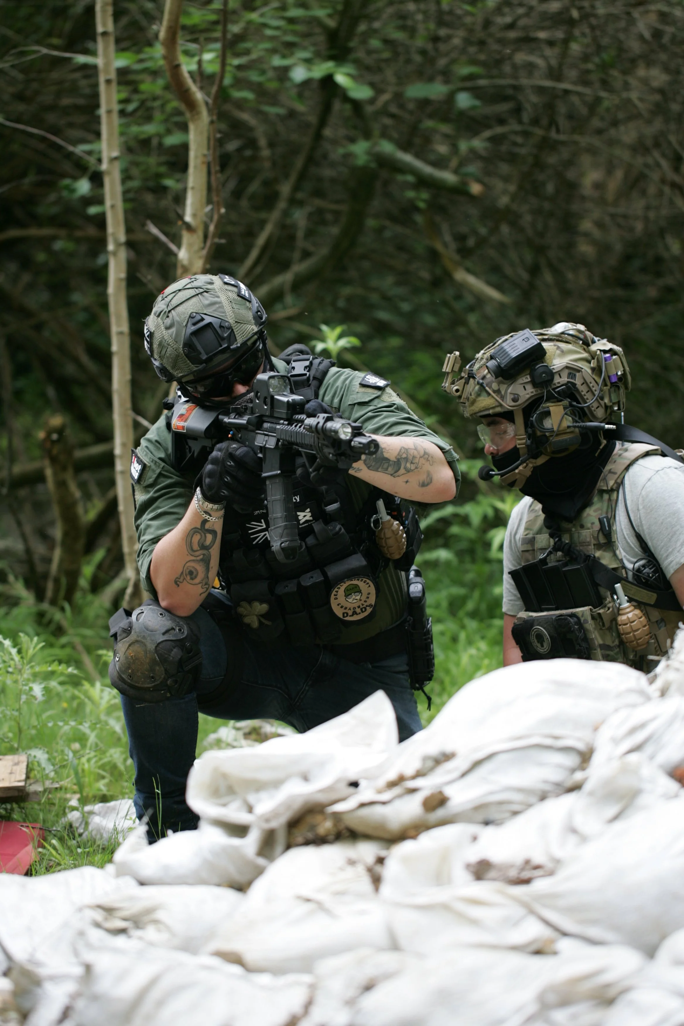 Two military personnel in tactical gear and helmets examining an object or area in a forested setting, with a pile of white fabric or material in the foreground.