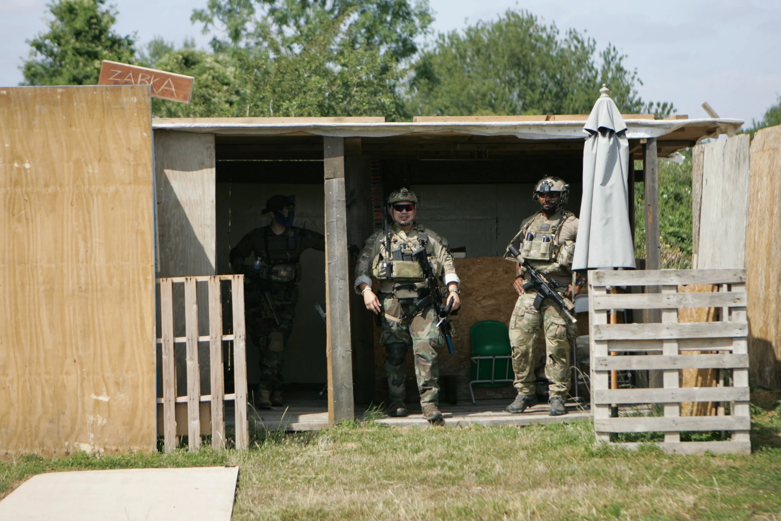Three soldiers in military gear standing at the entrance of a wooden structure, with a person inside wearing a mask and holding a water bottle.