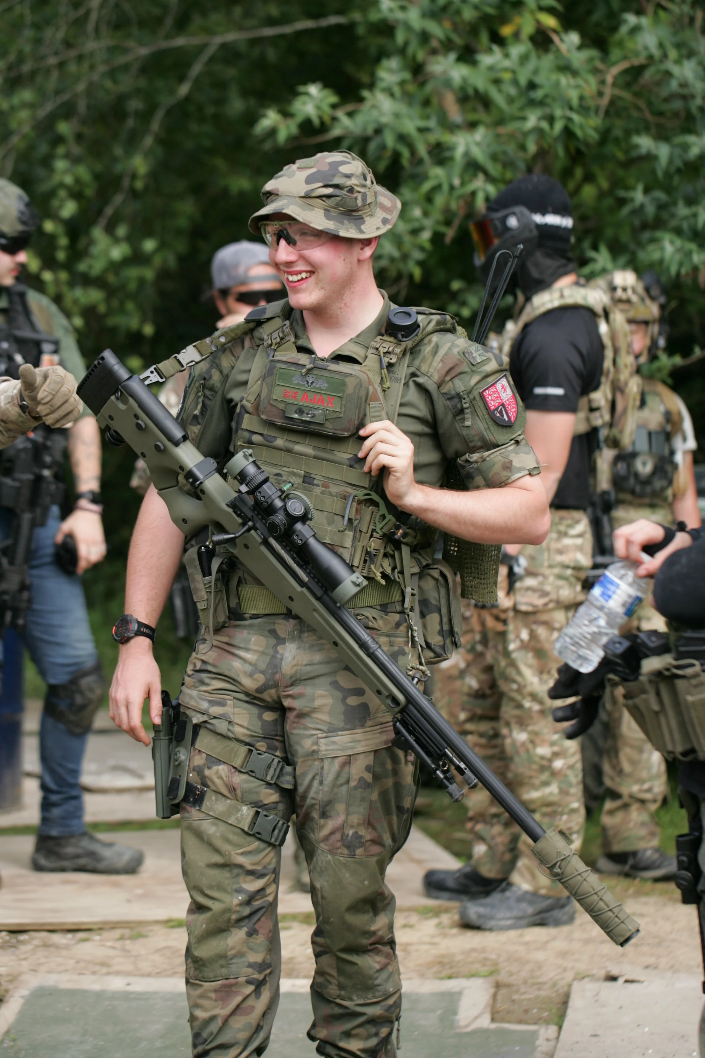 A group of soldiers in camouflage uniforms standing outdoors, with one soldier smiling and holding a large rifle in the foreground. The background has more soldiers and greenery.