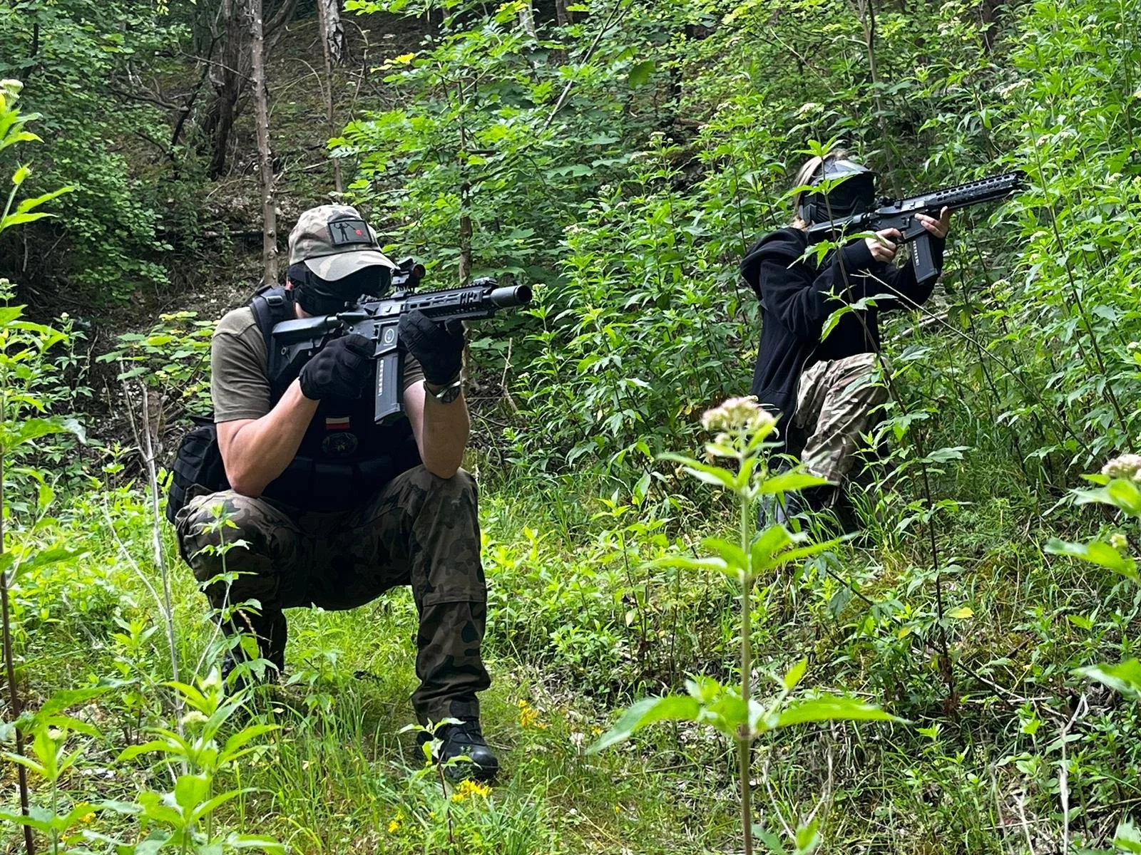 Two individuals wearing camouflage pants and tactical gear, using rifles in a wooded area.