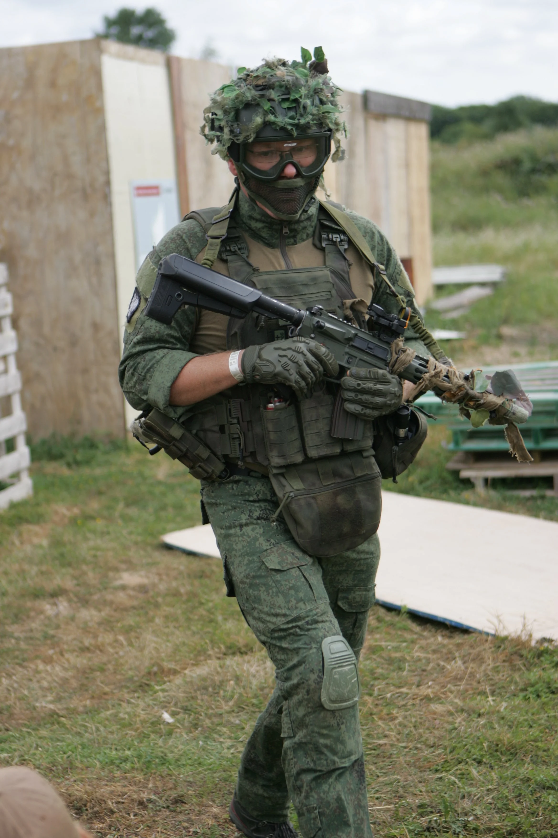 A soldier in camouflage uniform, wearing tactical gear and protective glasses, carrying a rifle, outdoors with wooden barriers in the background.