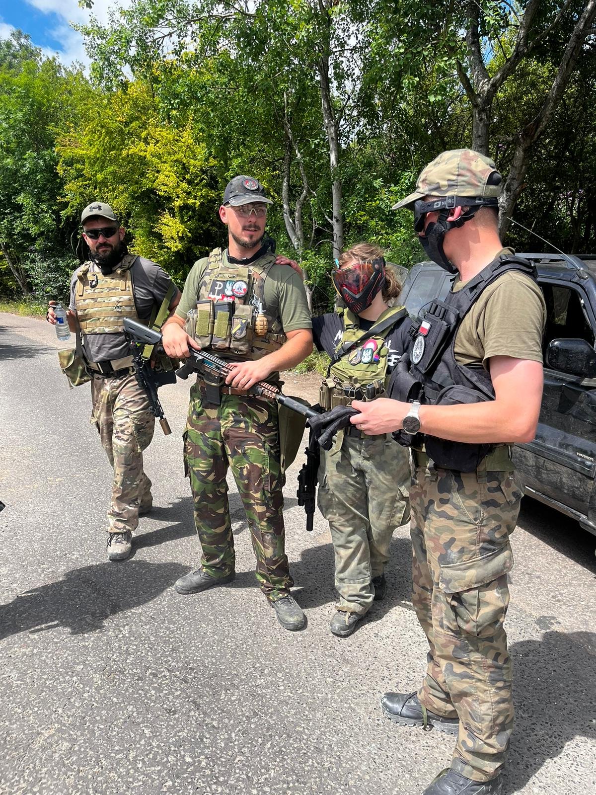 Four people dressed in military or tactical gear standing on a road near trees, engaged in conversation, with one person holding a water bottle and another holding a weapon.