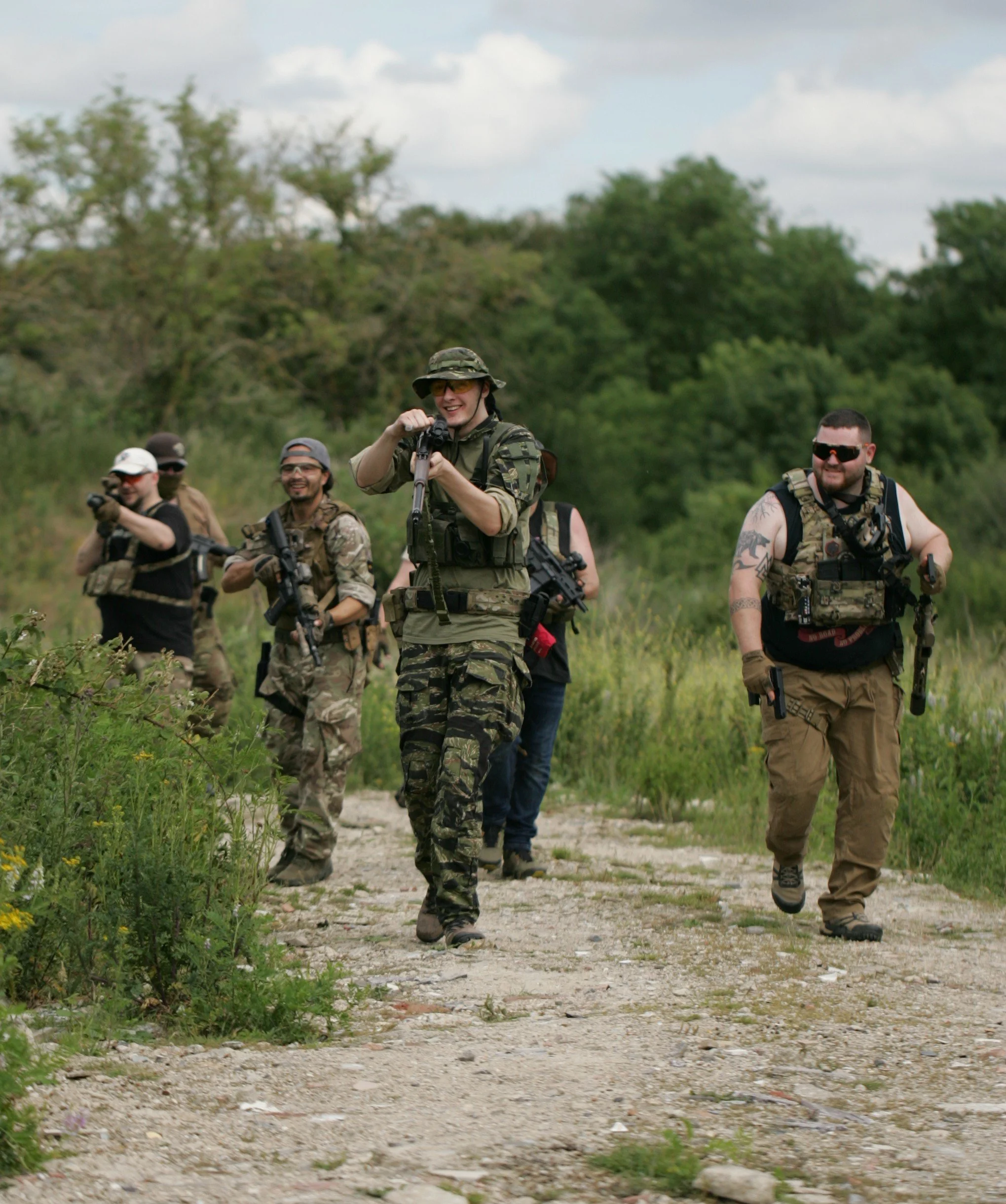 Group of soldiers in camouflage and tactical gear walking outdoors on a dirt trail in a green, wooded area.