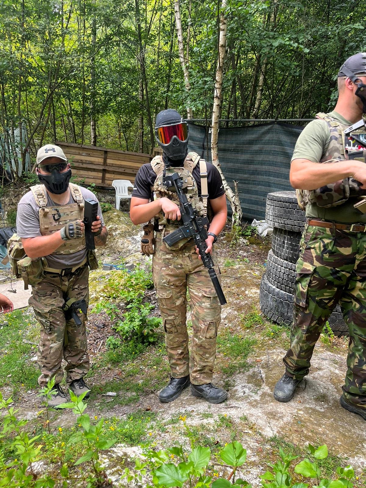 Three individuals in camouflage military clothing and gear standing outdoors in a wooded area, holding airsoft guns. They are wearing masks and protective eyewear, with one person in the center holding a rifle and the other two with smaller guns. The