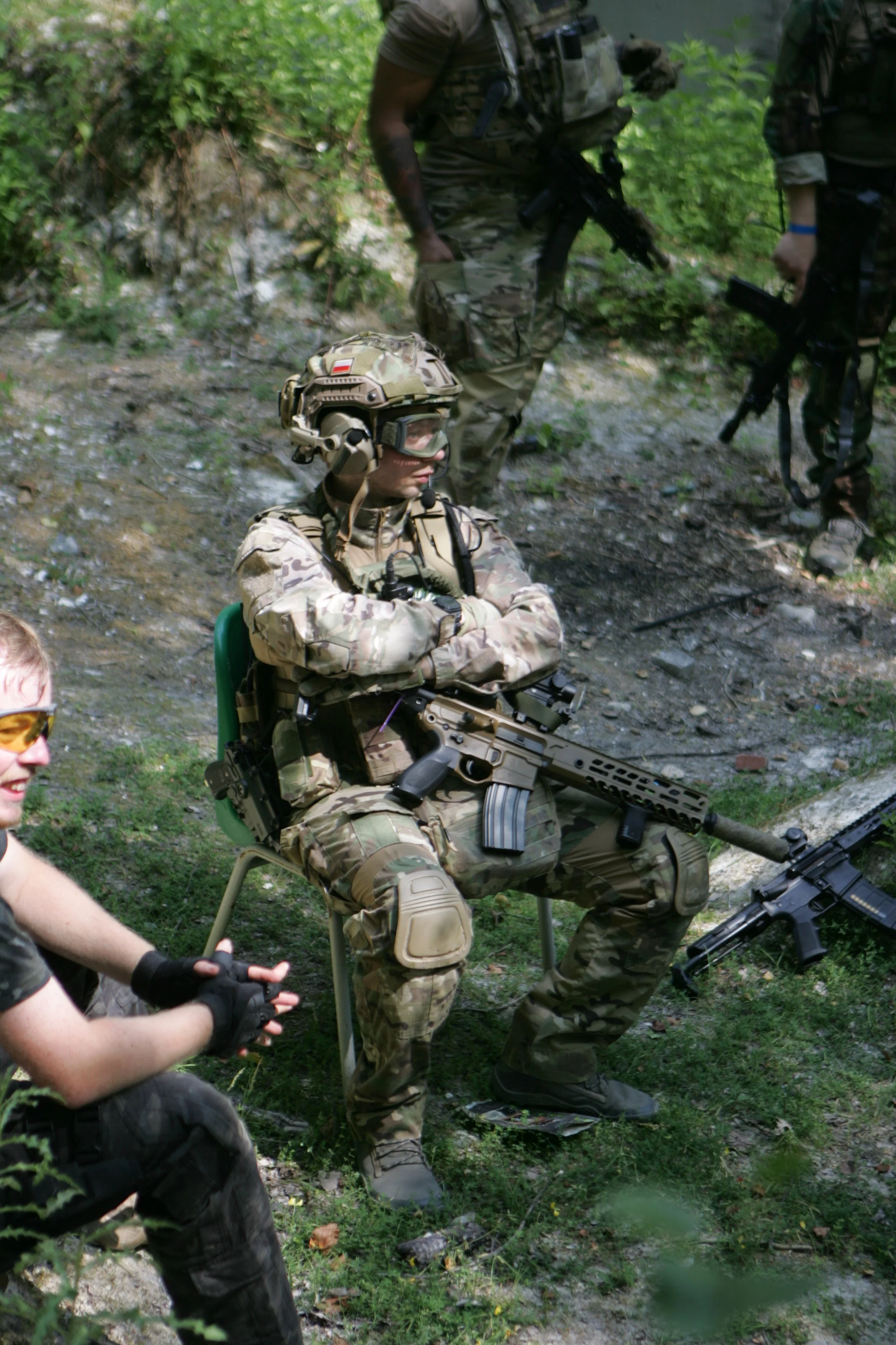 Soldiers in camouflage uniforms with guns in a forested area, one sitting and another standing, with a helmet and tactical gear.