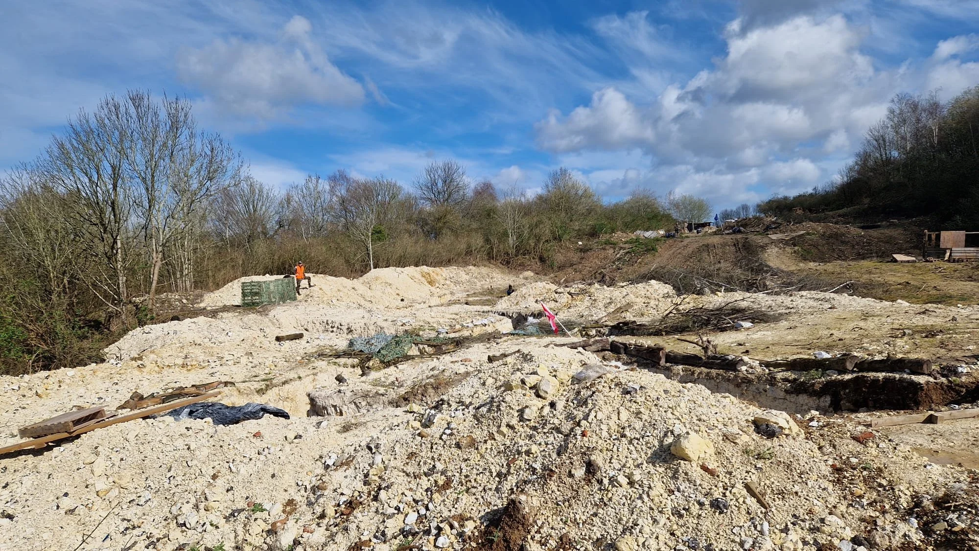 A construction site with dirt and debris, some fencing, and a worker in an orange safety vest. In the background, there are trees and a partly cloudy sky.