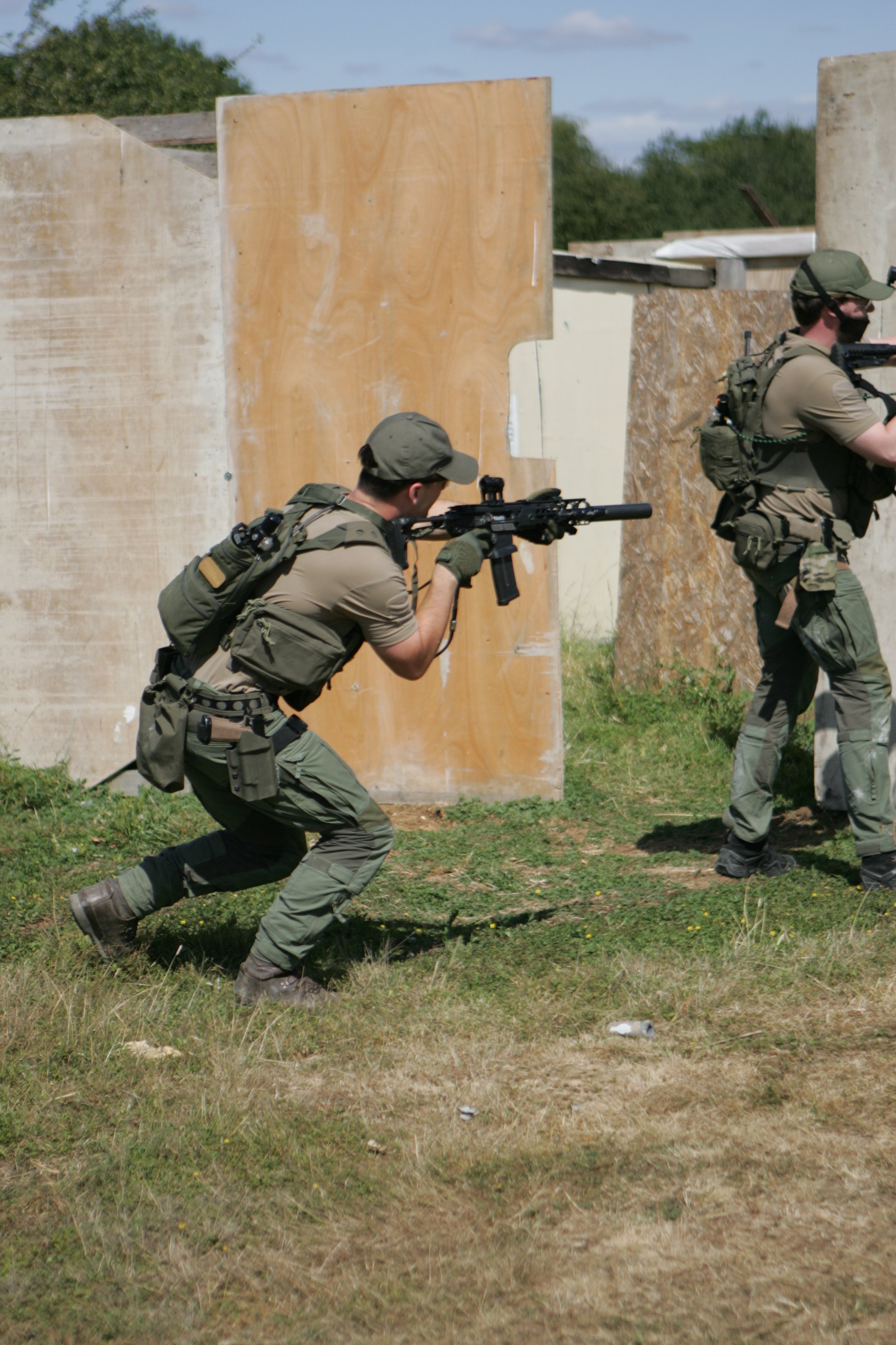 Two soldiers dressed in combat gear participating in a training exercise outdoors, with one kneeling and aiming a rifle while the other is standing behind a wall with a rifle.