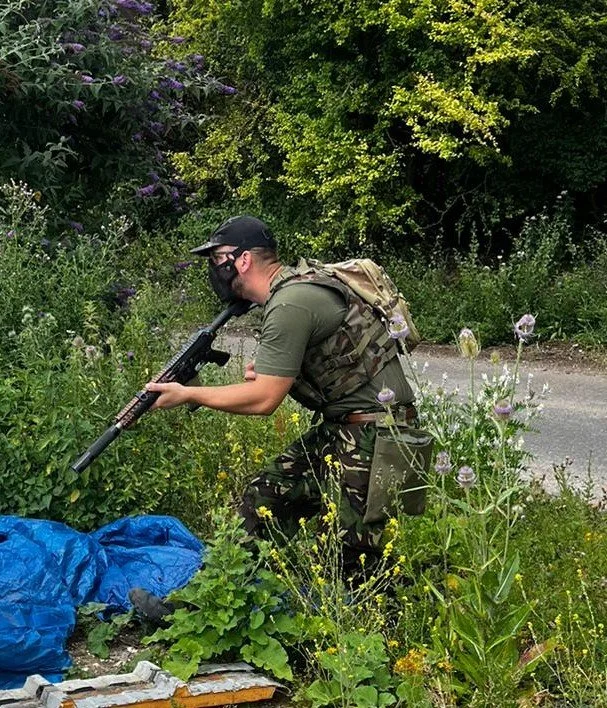 A man in military camouflage uniform, wearing a black mask and black cap, is crouching among green plants and wildflowers by the side of a paved road, holding a rifle.
