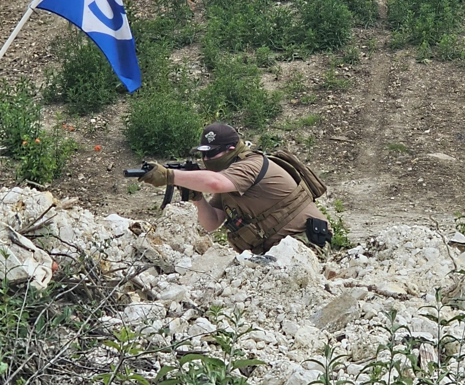 A person wearing military or tactical gear, aiming a rifle while crawling on rocky terrain.