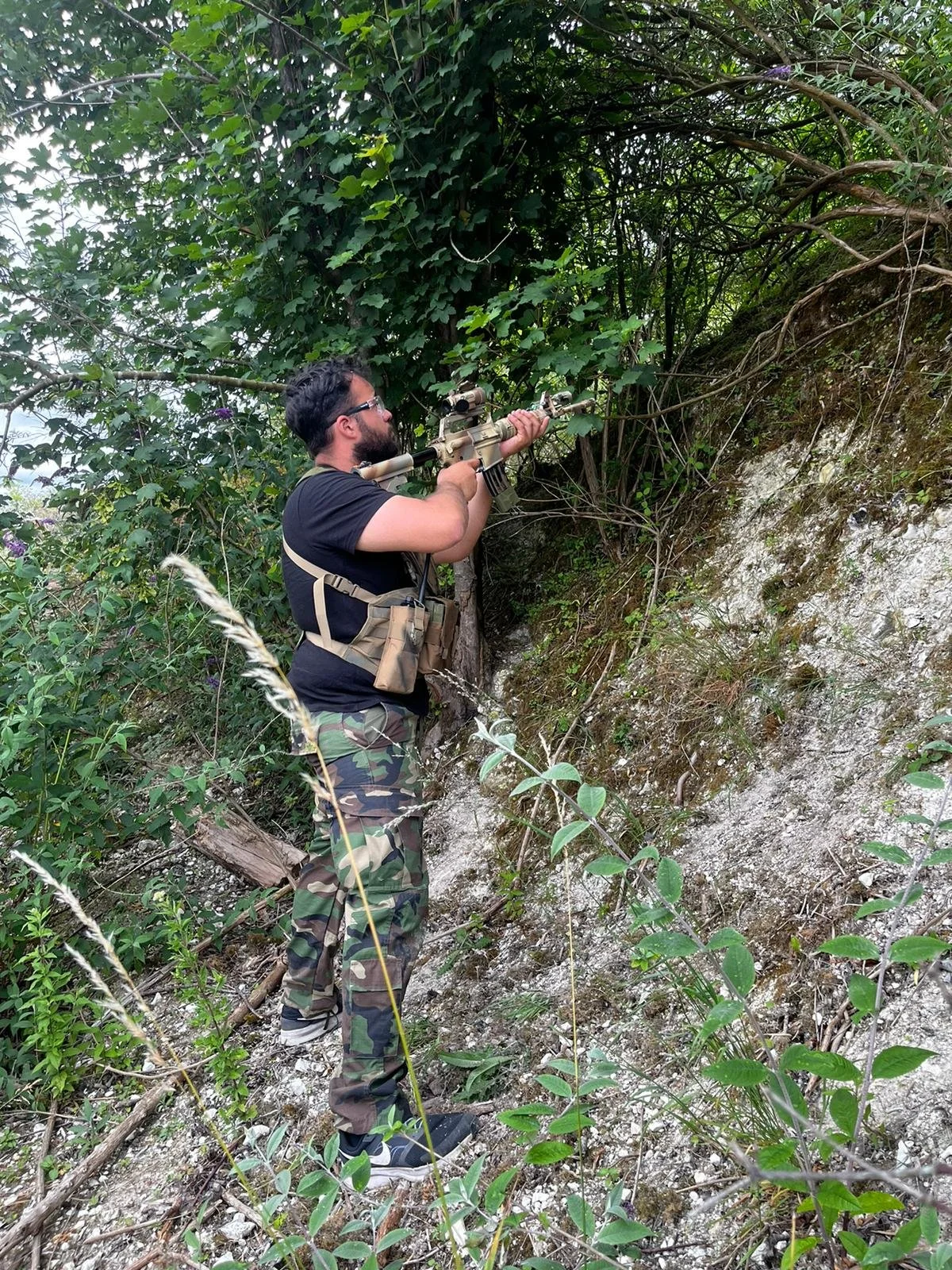 Man aiming a sniper rifle while standing on a hillside with dense greenery and exposed soil.