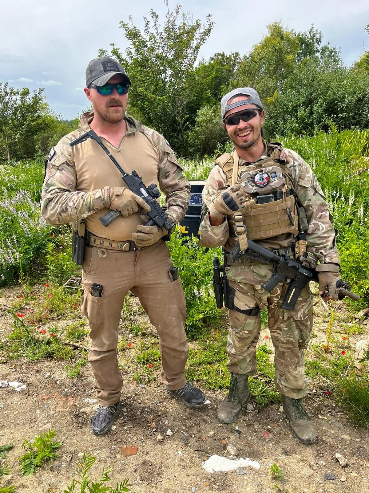 Two men in military or tactical gear standing outdoors in a grassy and wooded area, holding rifles, wearing sunglasses, and smiling or looking serious.