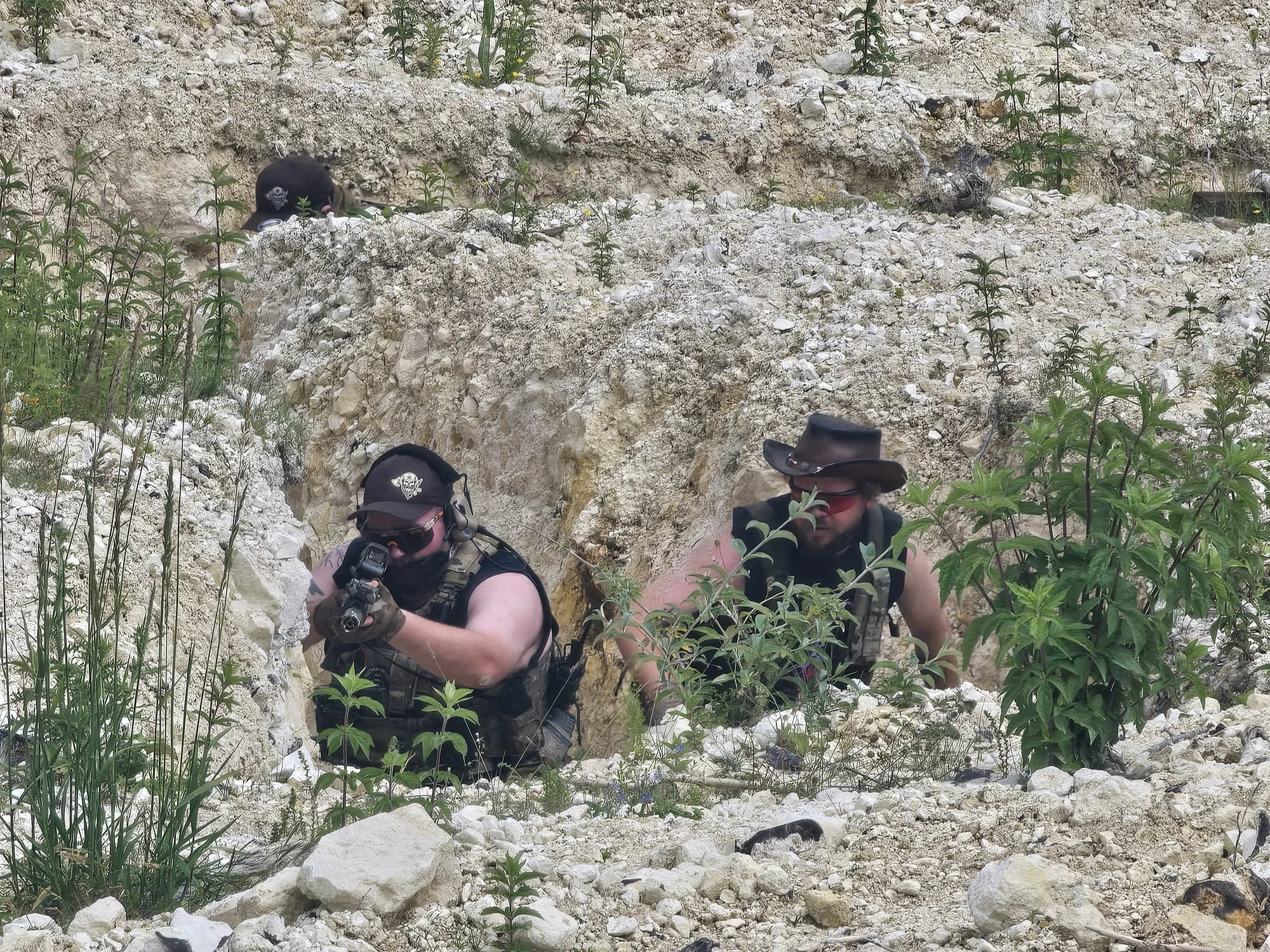 Two people wearing tactical gear and headphones, holding guns, hiding among rocks and green plants in a rocky outdoor environment.