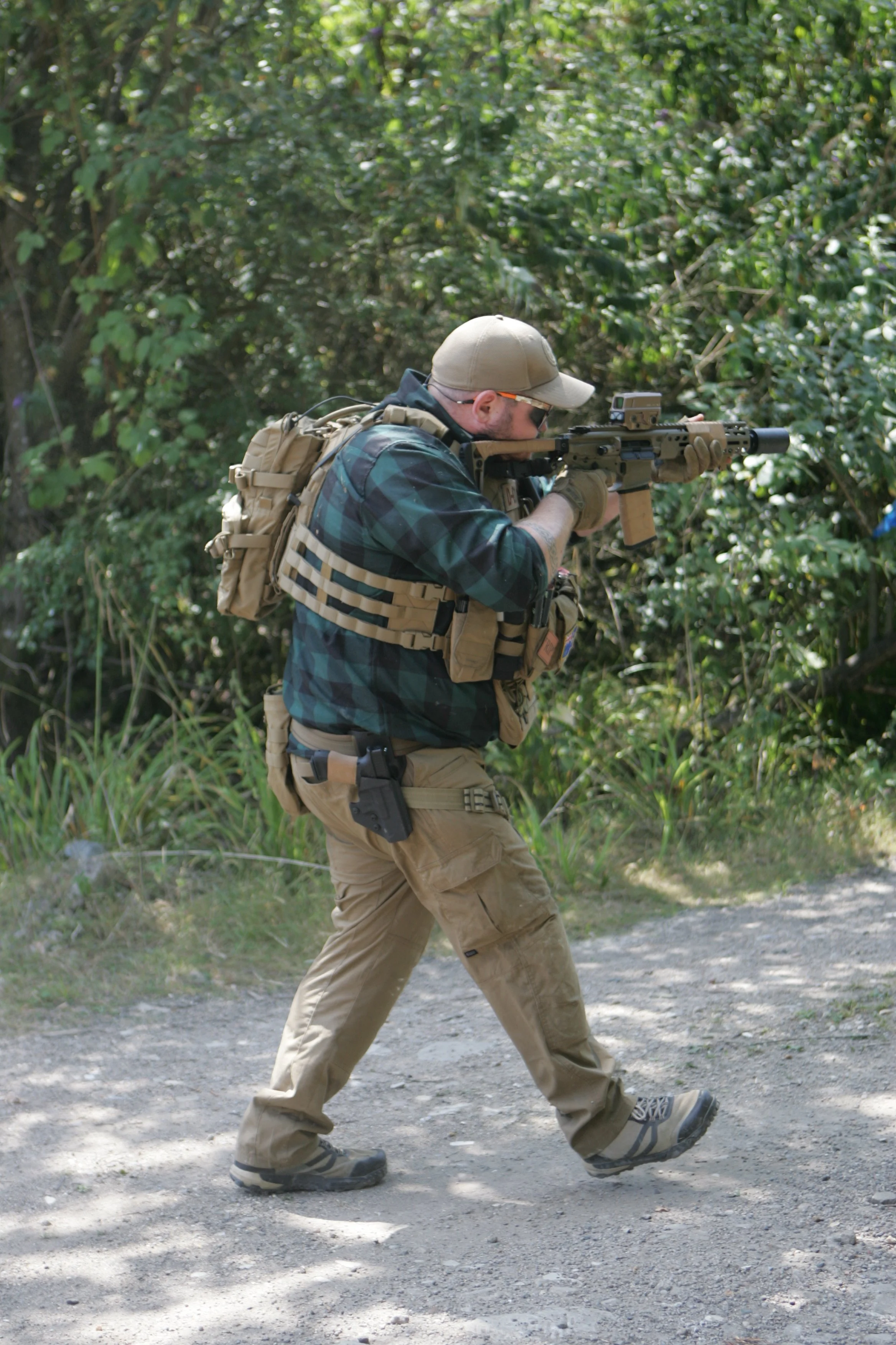 A man in tactical gear aiming a rifle outdoors in a wooded area.