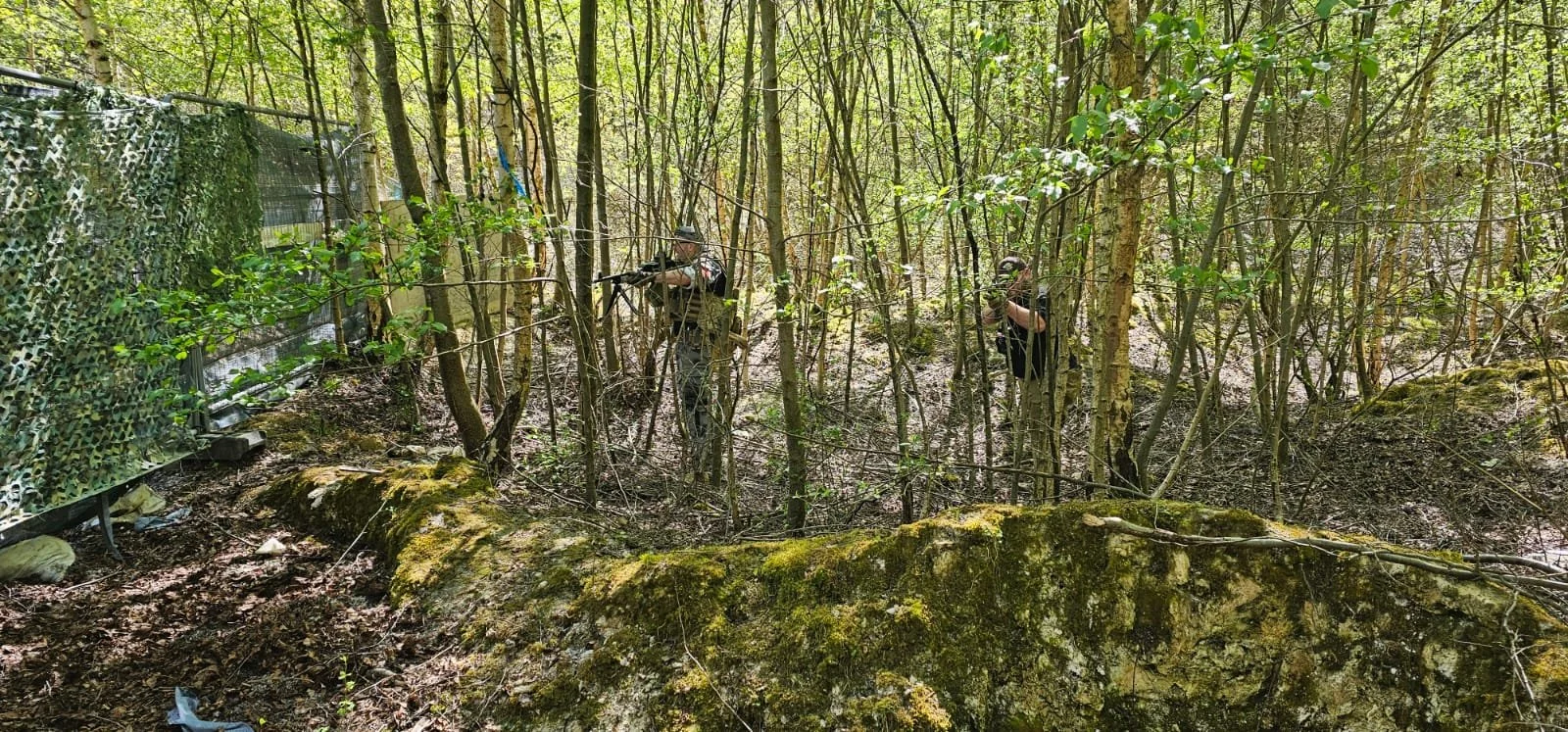 Two armed individuals wearing tactical gear and helmets in a dense wooded area, surrounded by trees and moss-covered logs.