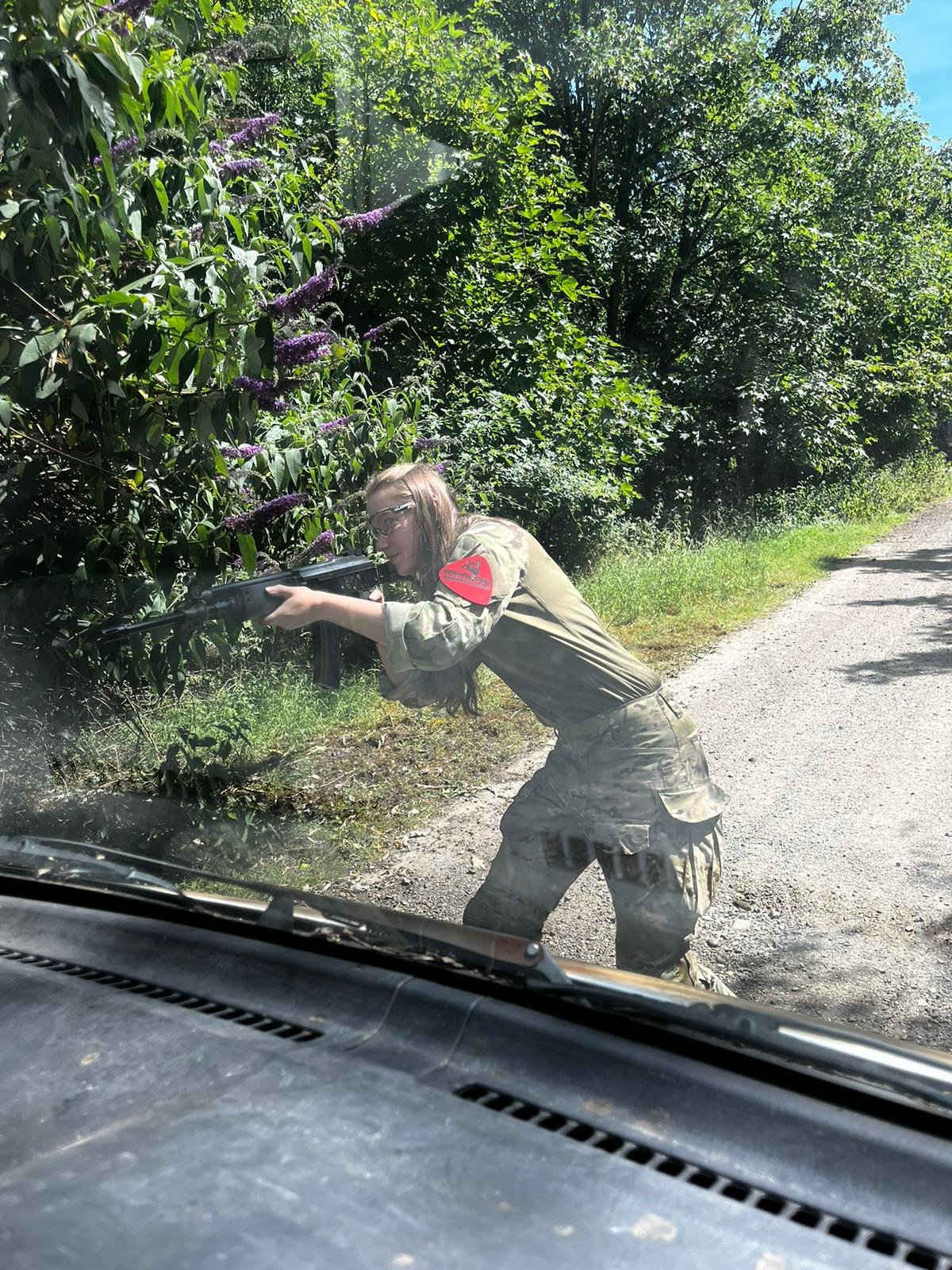 A person wearing military camouflage pants and a beige shirt aiming a rifle near the bushes by the side of the road, viewed through a car windshield.