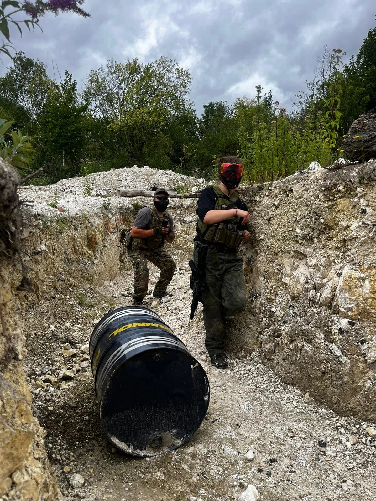 Two individuals dressed in tactical gear and camouflage clothing are in a rugged outdoor area with dirt and rocks, possibly on a military or paintball mission. One person in the foreground has a red-tinted mask and a tactical vest, while the other in