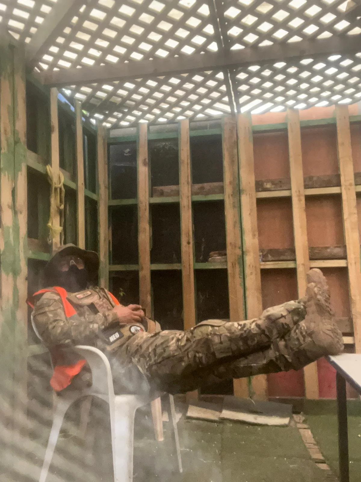A person in camouflage military fatigues with a black face mask, wearing that has an orange safety vest, reclines in a white plastic chair inside a wood-framed unfinished room. The person appears to be using a mobile phone, with one leg crossed over 
