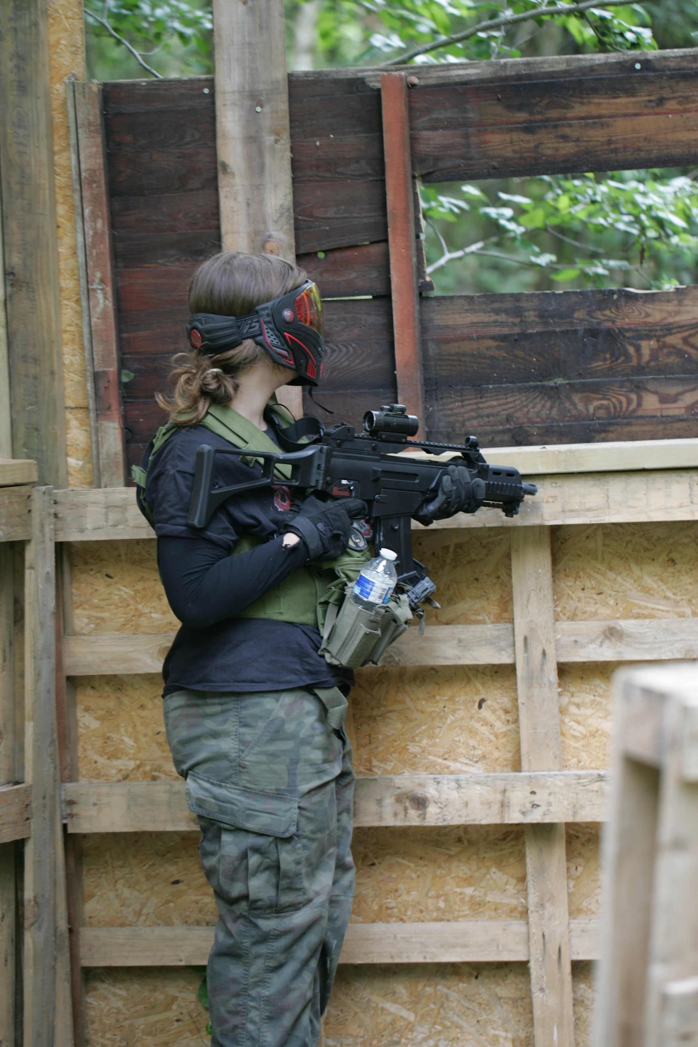 A woman holding a paintball gun, wearing a protective mask and camouflage pants, stands inside a wooden structure with a partial wall, surrounded by trees.
