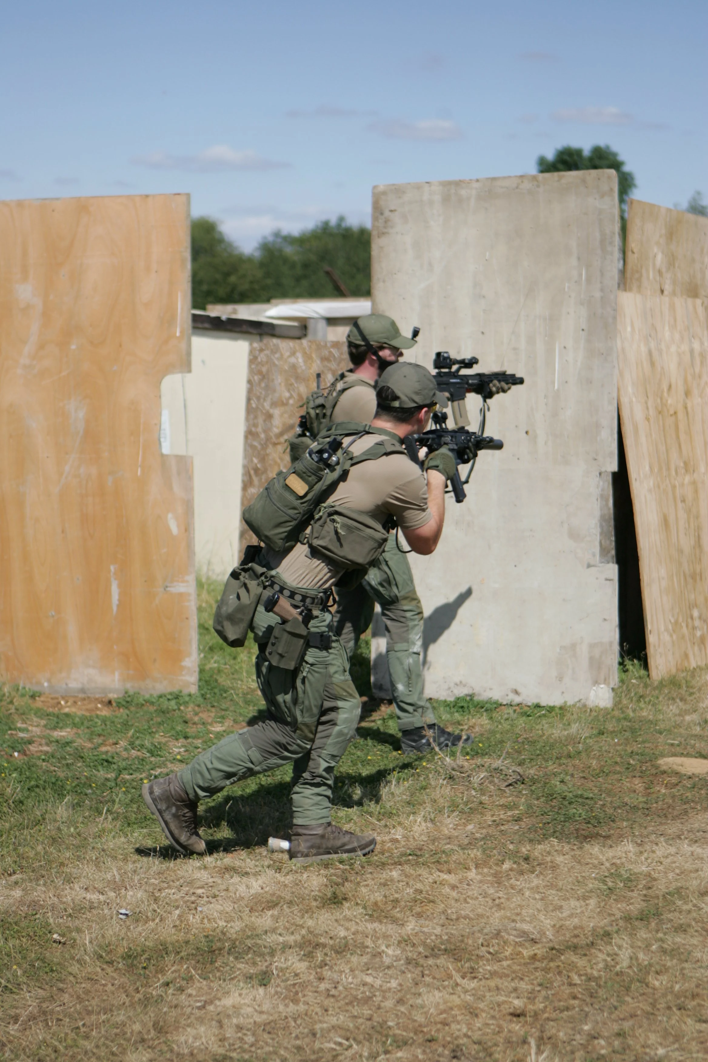 Two soldiers in military gear advancing through a makeshift obstacle course made of wooden panels, outdoors on grassy terrain, during daytime.