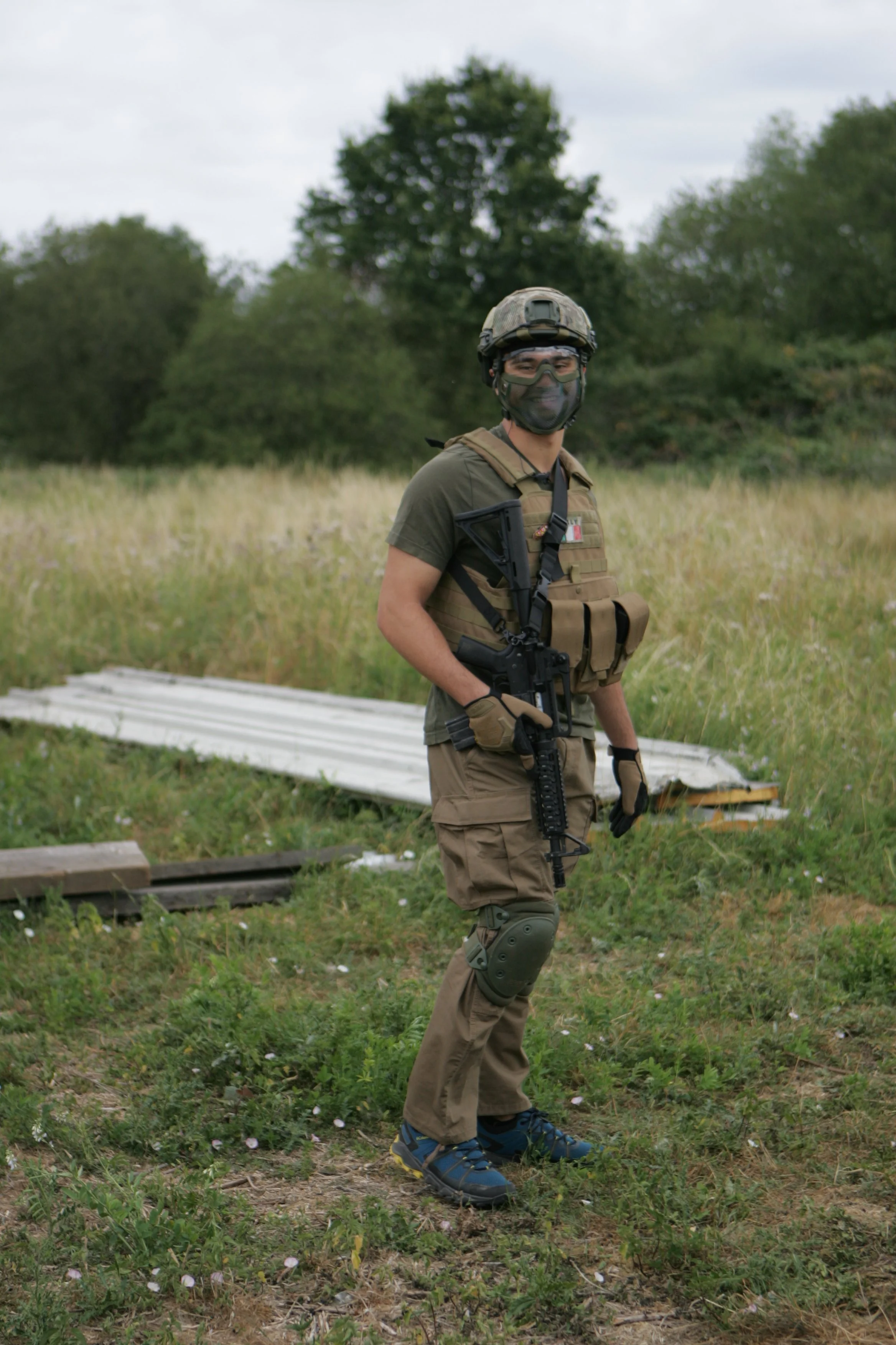 A soldier or military personnel dressed in tactical gear, including a helmet, face mask, vest, and knee pads, stands outdoors in a grassy area with trees and cloudy sky in the background.