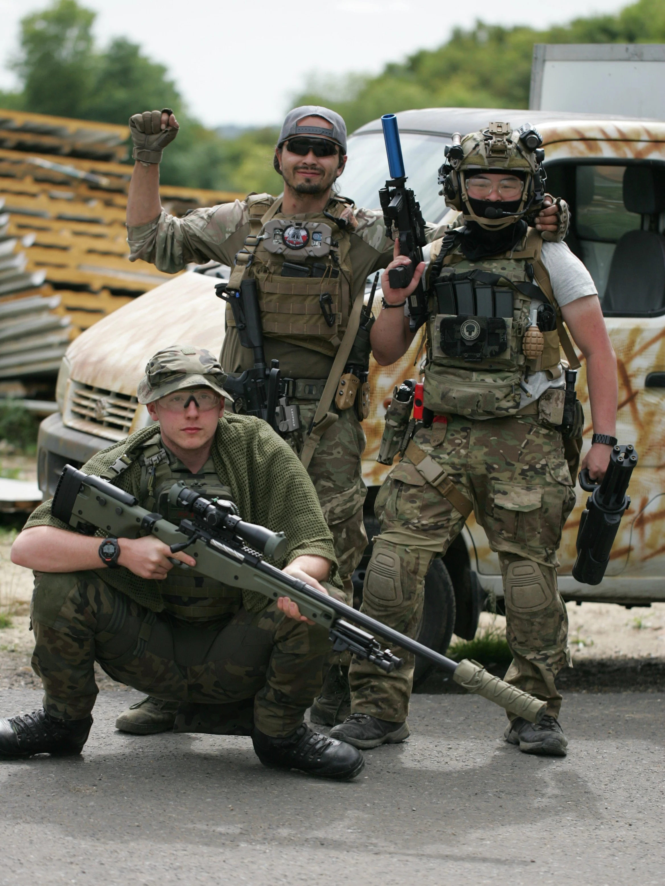 Three military personnel in camouflage uniforms with gear, weapons, and protective gear in front of a rusted vehicle and a wooden stack outdoors.