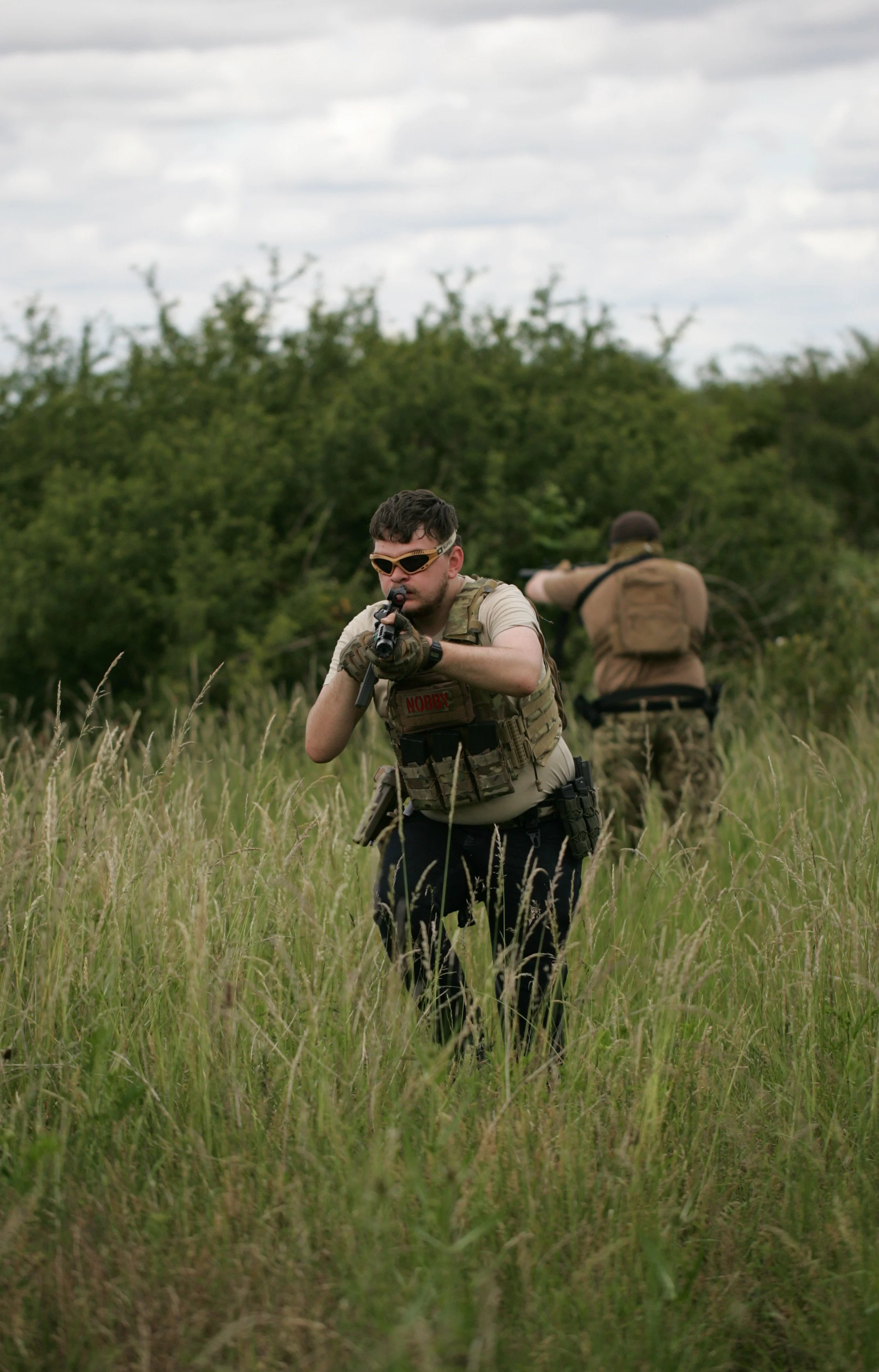 Two men in camouflage and tactical gear aiming firearms in a grassy field with trees in the background.