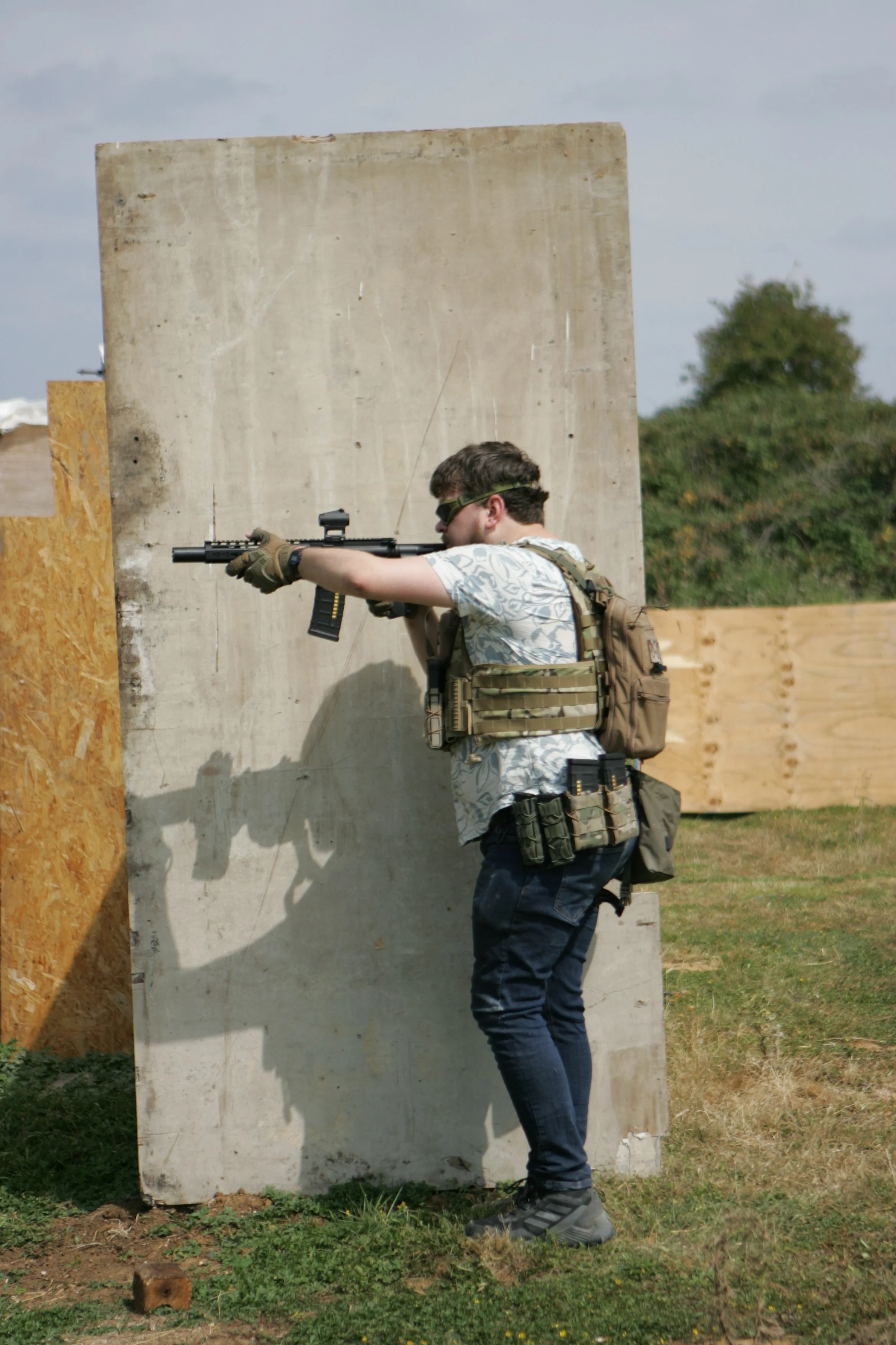 Man with tactical gear aiming a rifle around a concrete wall at an outdoor shooting range.