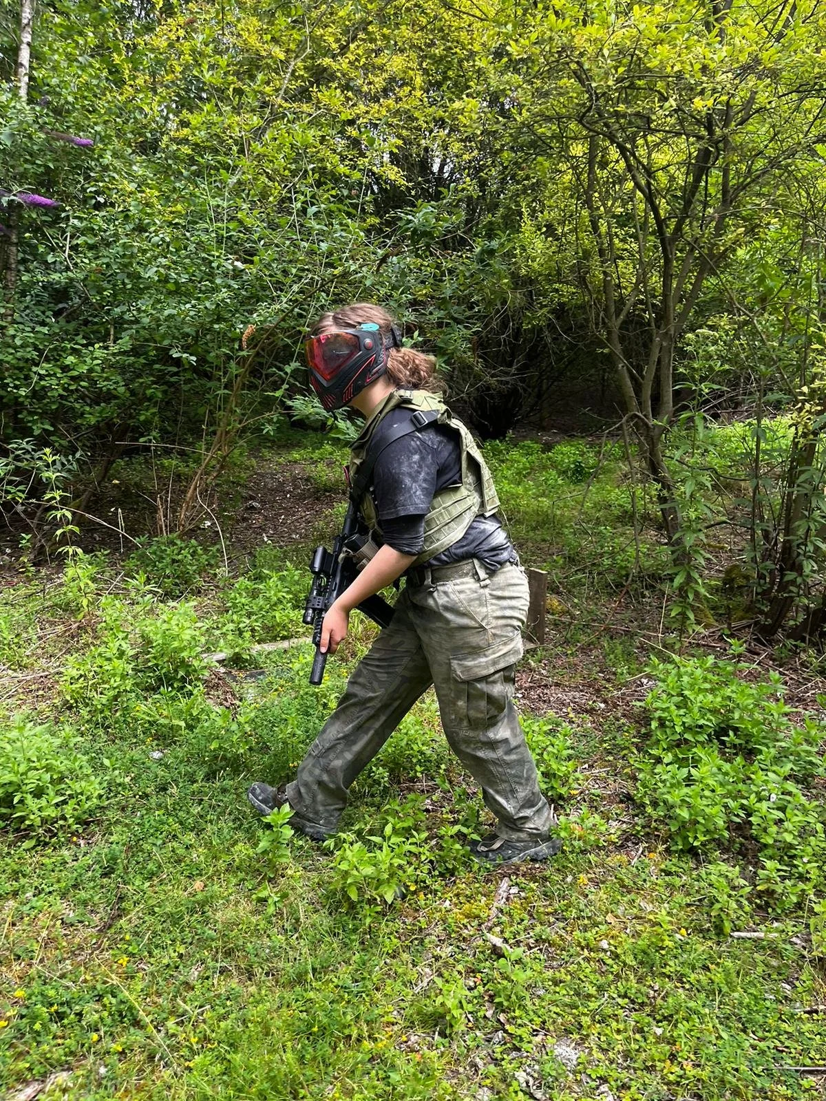 A person in military camouflage pants wearing a vest and a helmet with a visor, carrying a paintball gun, walking through a wooded area with green bushes and trees.