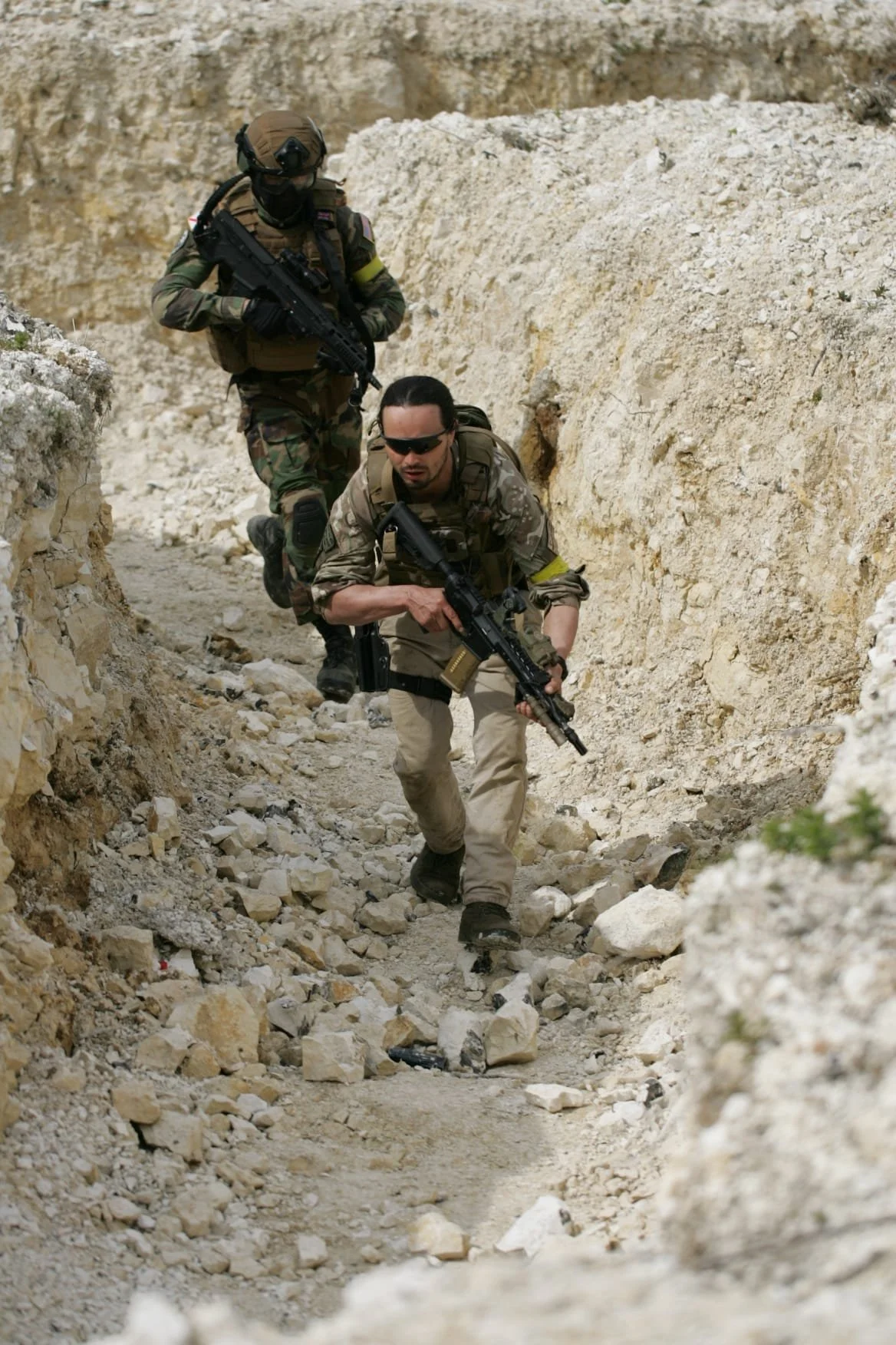 Two soldiers moving through rocky, desert terrain with weapons, dressed in tactical military gear and camouflage clothing.