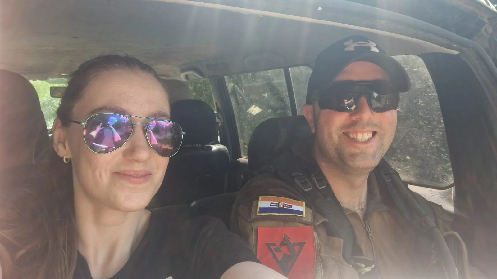 A man and woman smiling inside a vehicle, both wearing sunglasses. The man is in military or tactical gear, with patches on his uniform, including a flag patch and a red and black emblem, and is wearing a cap and sunglasses. The woman is wearing refl