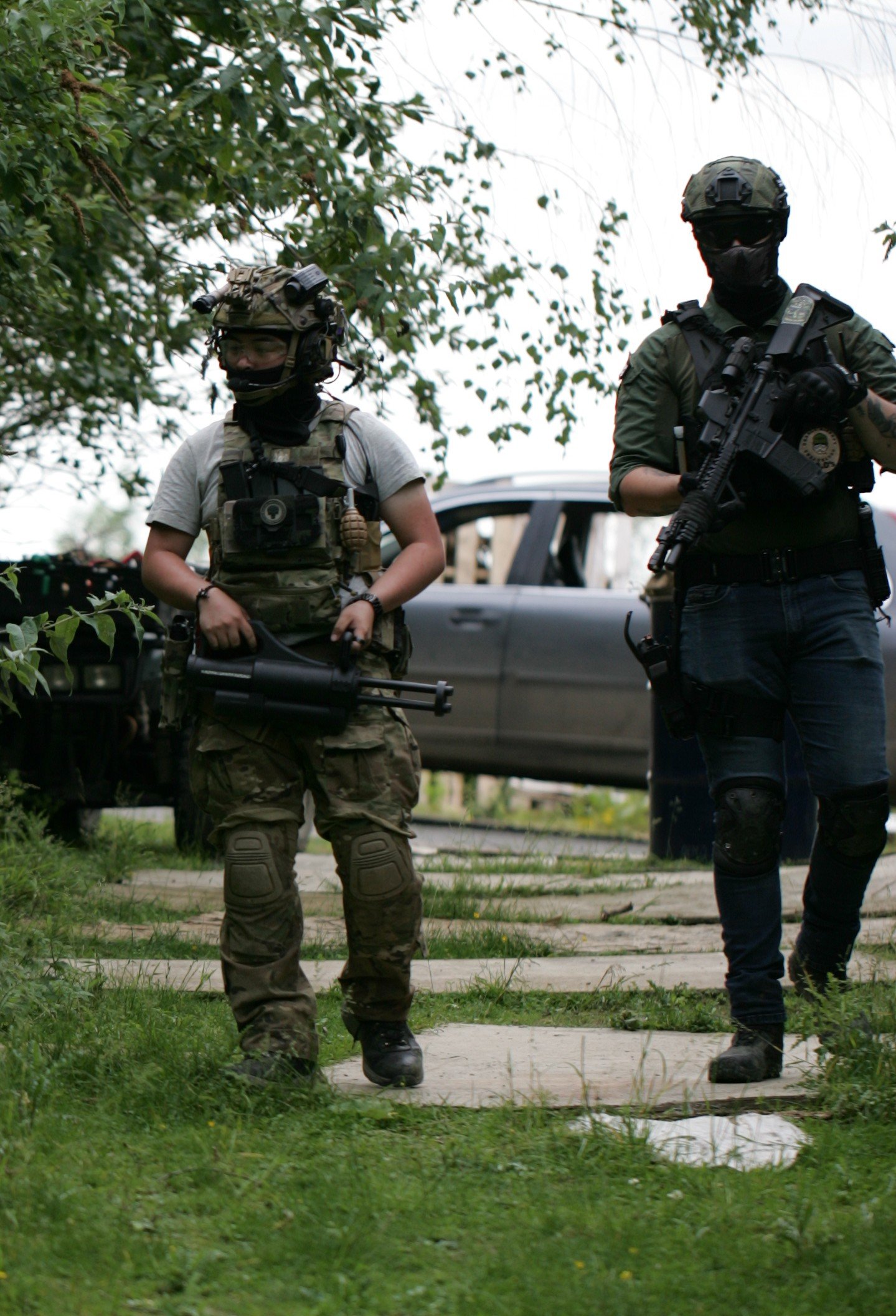 Two armed military personnel in tactical gear standing on a sidewalk, with greenery and a parked car in the background.