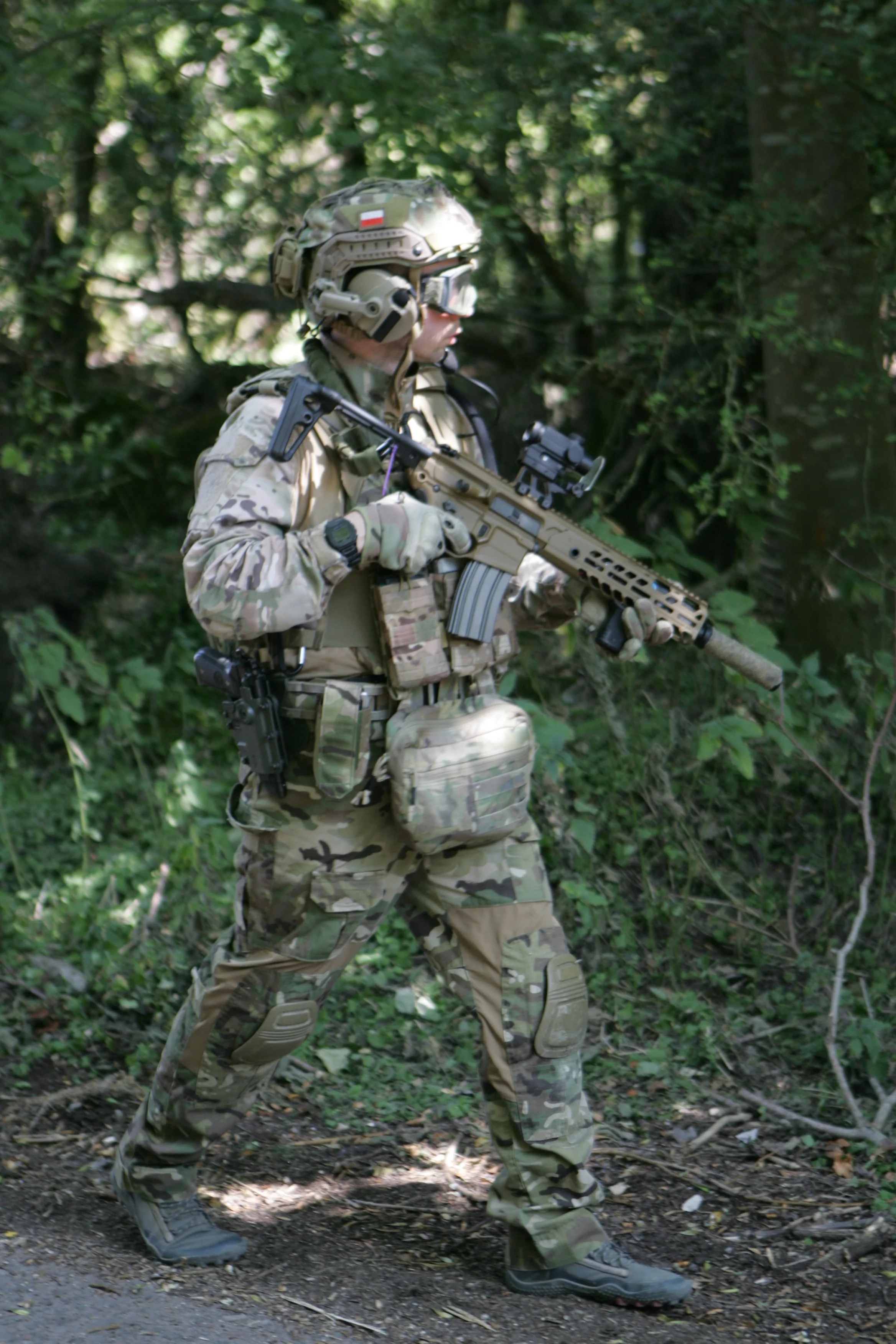 A soldier in camouflage military uniform walking through a wooded area while holding a rifle.