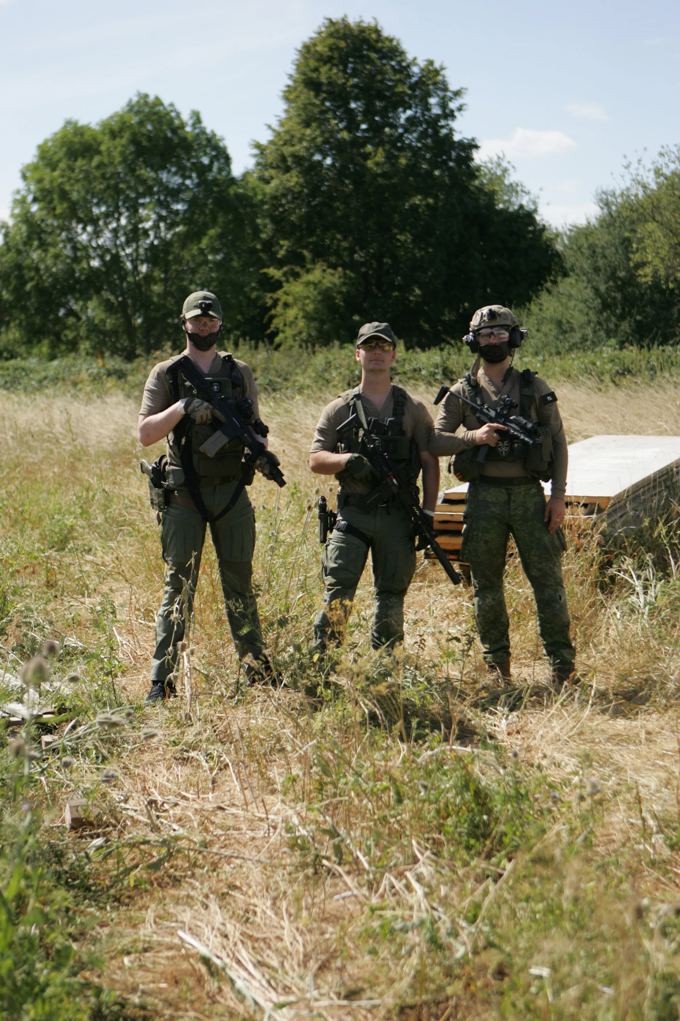 Three soldiers standing outdoors in a grassy field with trees and a blue sky in the background, holding rifles and wearing tactical gear.