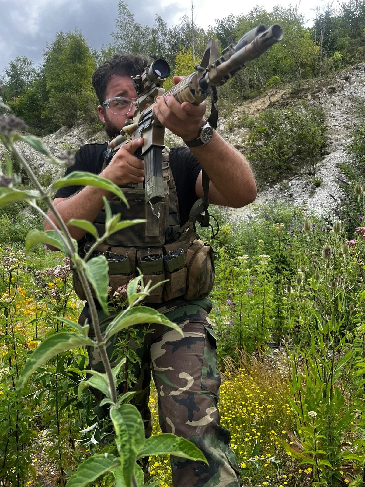 A man with a beard and glasses aiming a scoped rifle during daytime outdoors in a green, wooded area with wildflowers and a rocky hillside.
