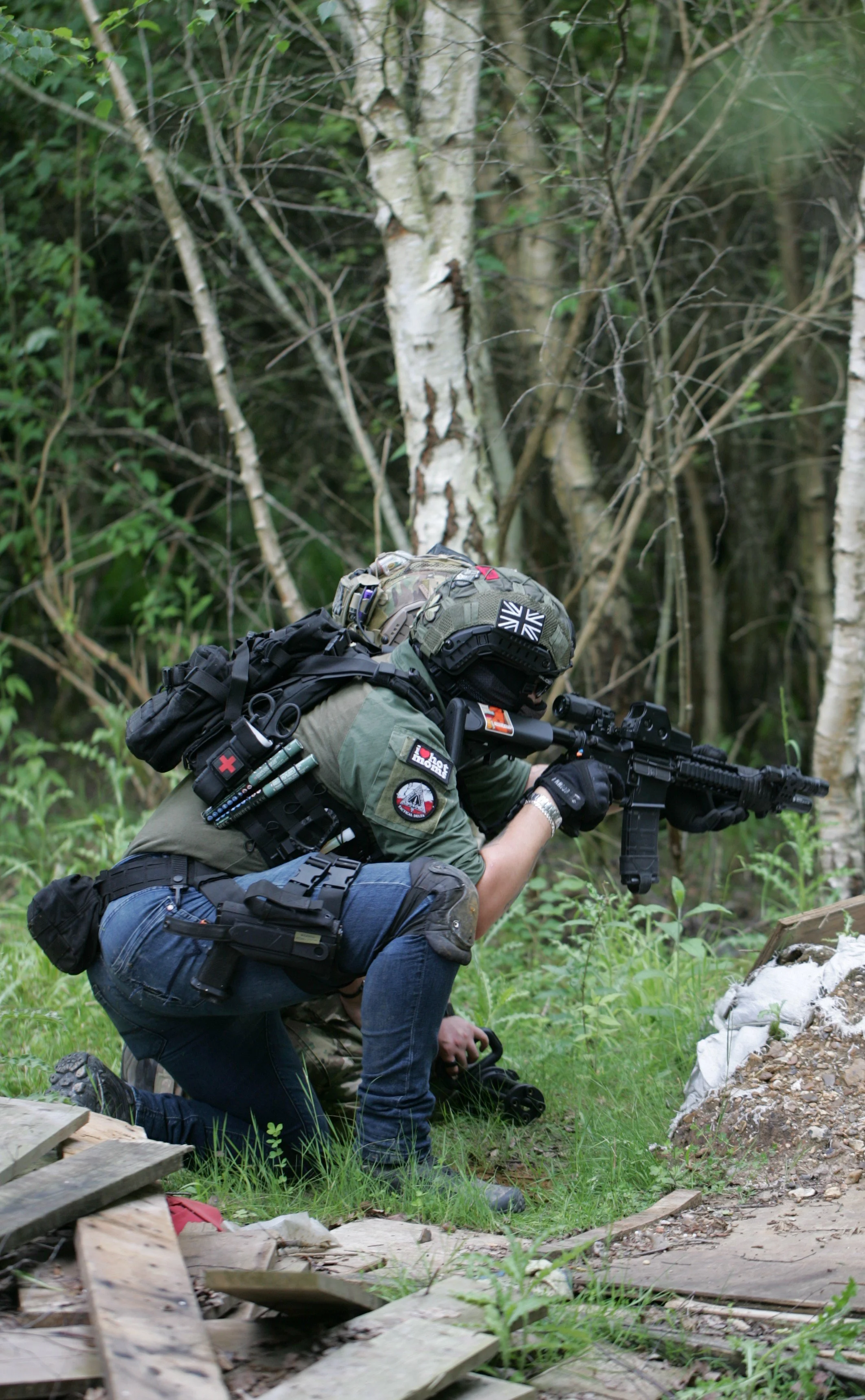 A soldier wearing camouflage gear and a Union Jack patch, kneeling and aiming a rifle in a forested area.