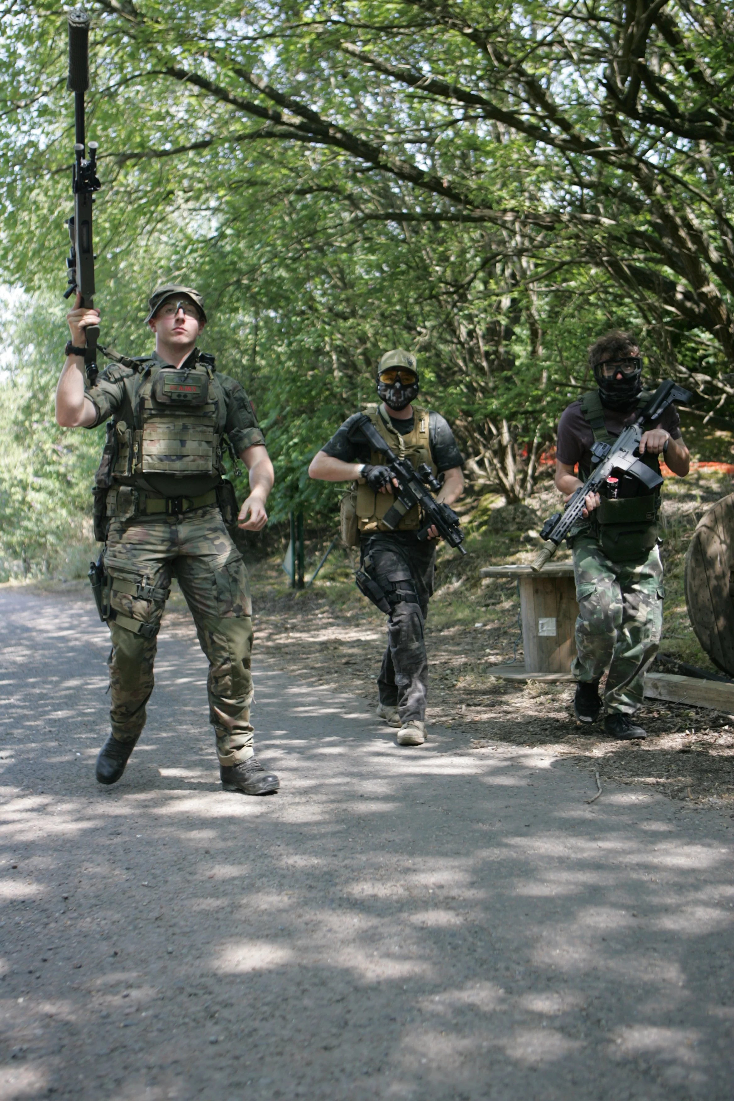Three people dressed in tactical military gear, holding rifles, walking outdoors in a wooded area with green trees and a gravel path.