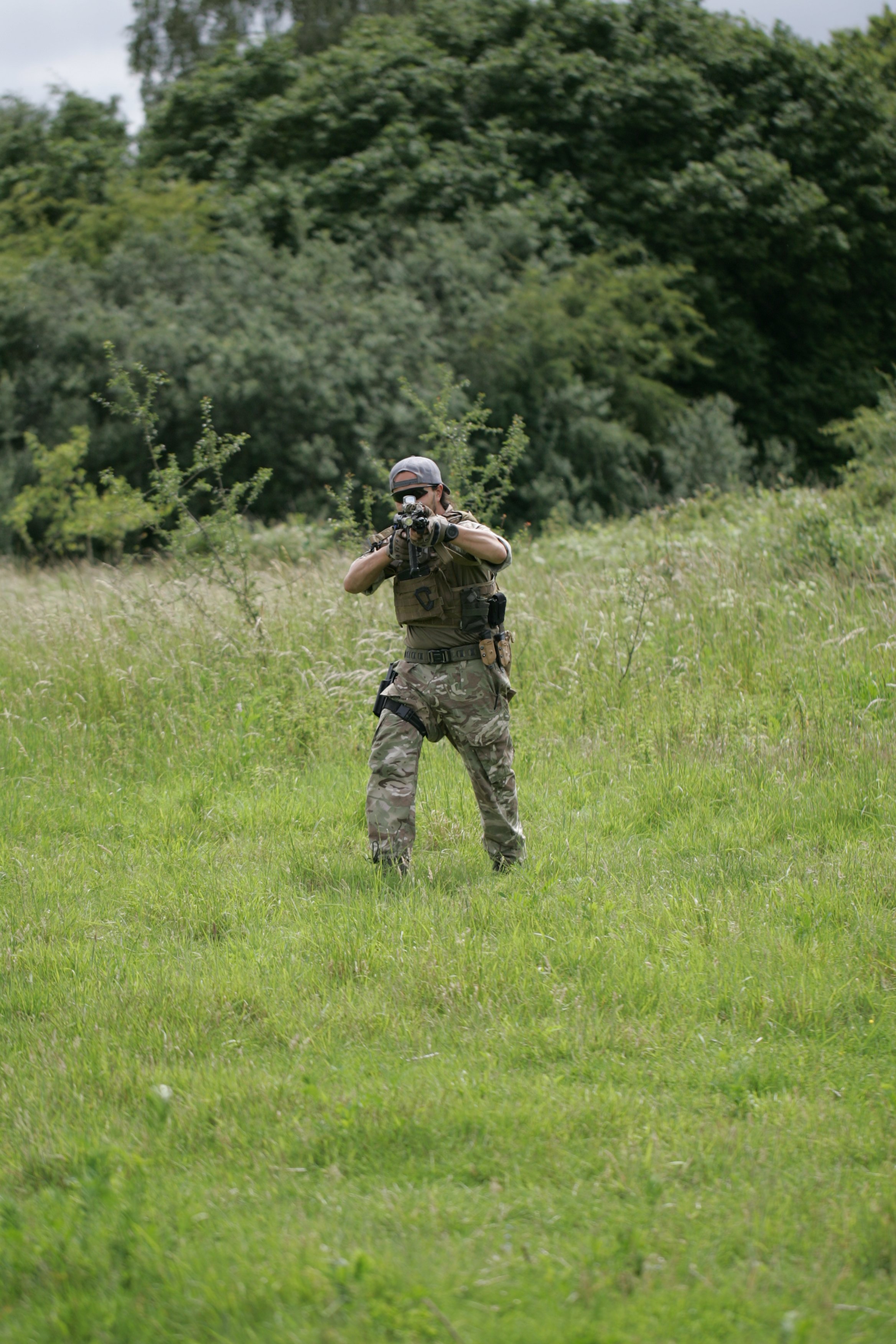 A person dressed in military camouflage pants and a tan shirt aiming a rifle while standing in a grassy field with trees in the background.