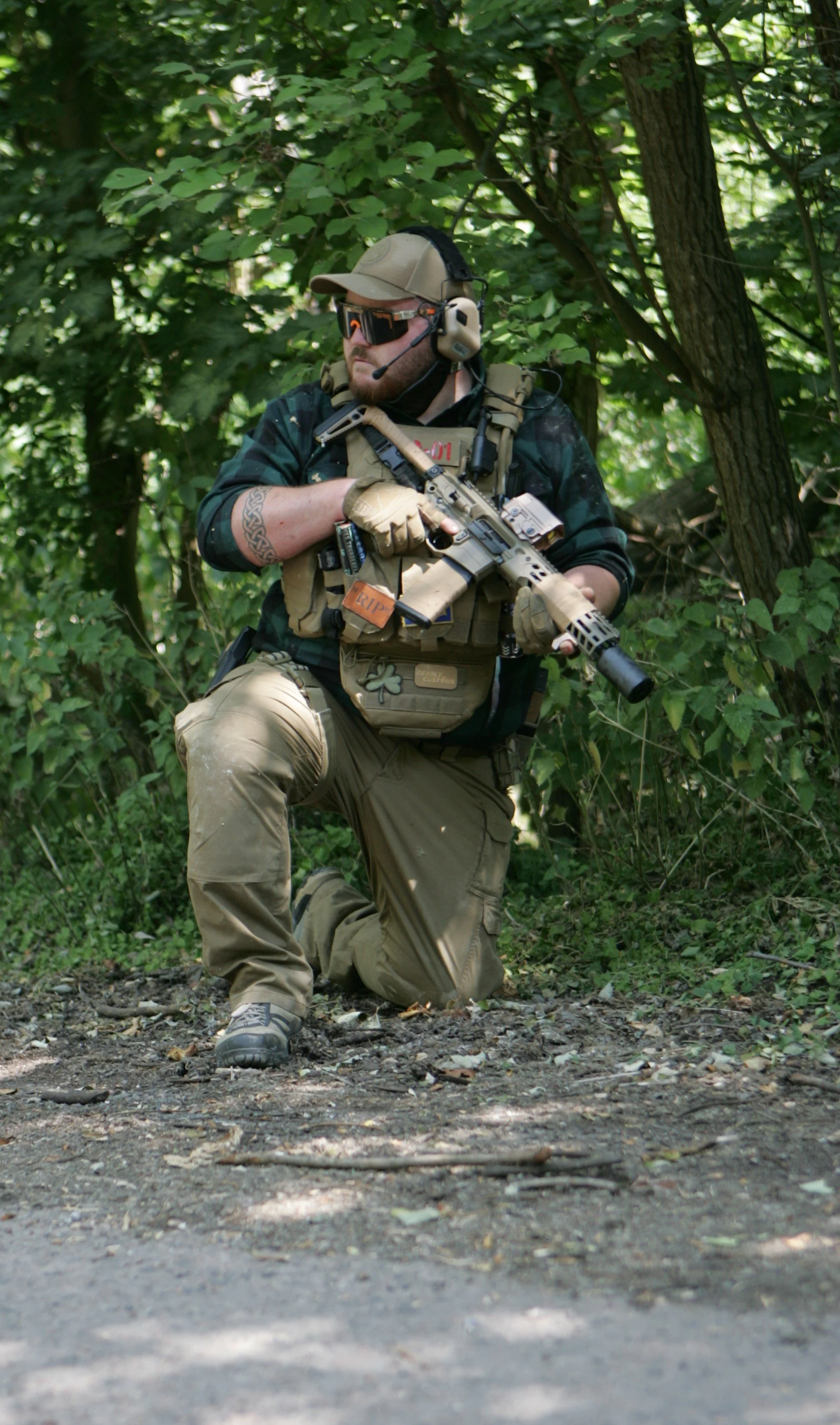 A man in tactical gear kneeling in a wooded area, holding a rifle, wearing sunglasses, a beige cap, and a headset with a microphone.