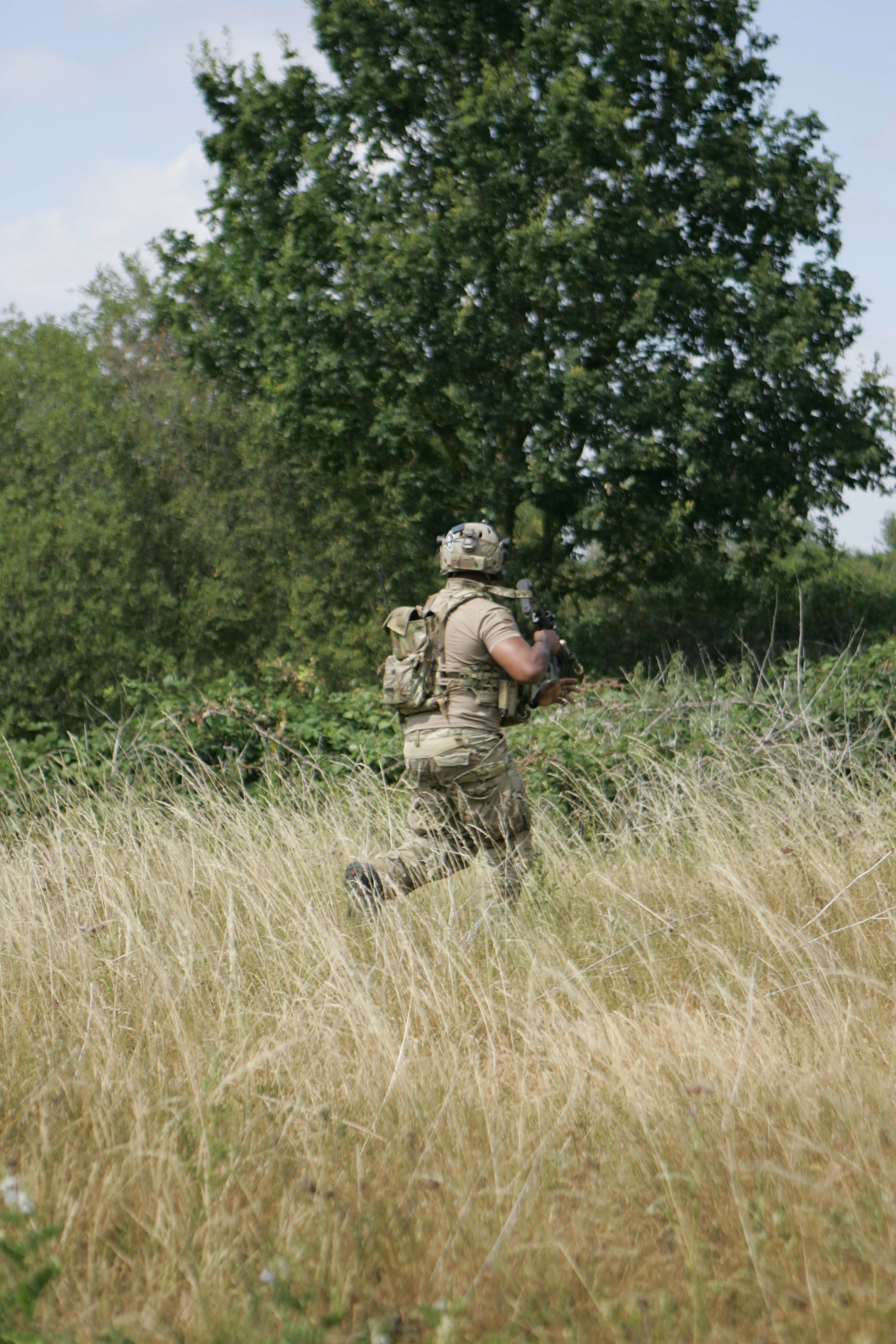 A soldier in camouflage clothing and a helmet running through a field of tall grass, with trees and a blue sky in the background.