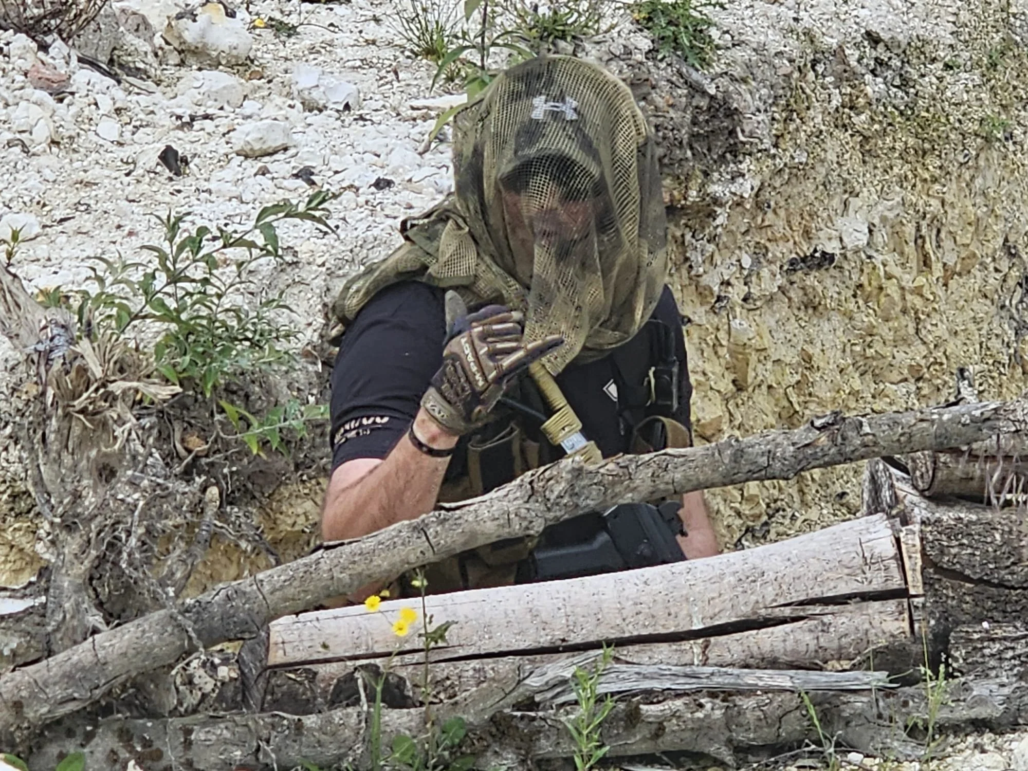 A person wearing a protective mask and gloves sitting among logs and rocks outdoors.