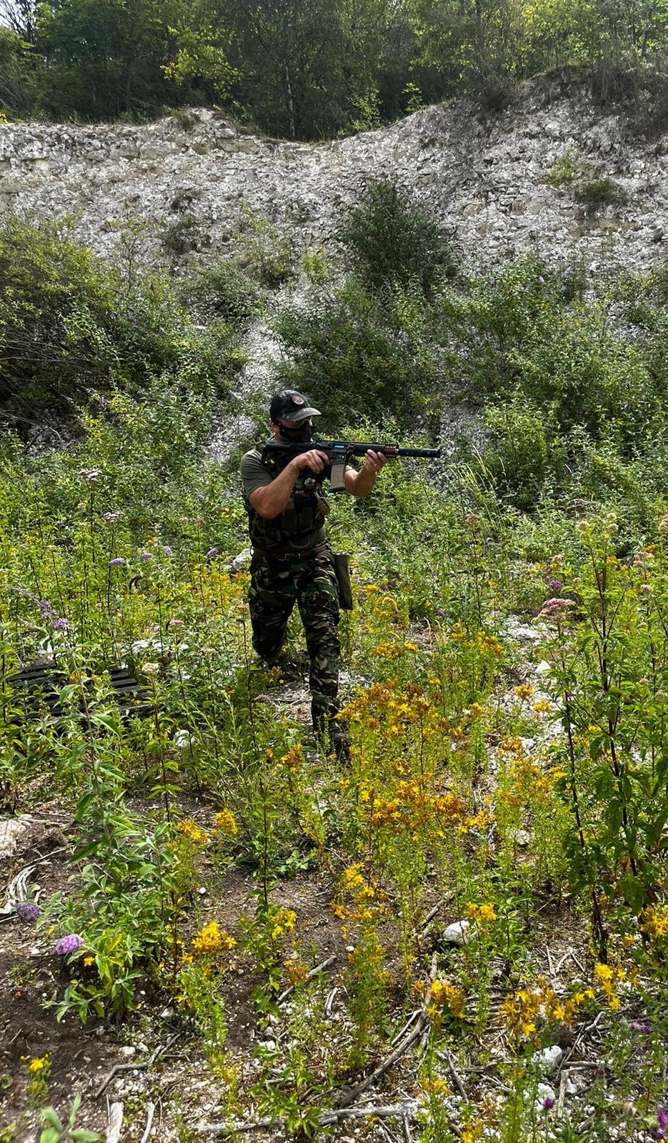 A soldier in camouflage clothing and tactical gear holding a rifle, walking through a field of tall green plants and yellow flowers with a rocky hillside and green trees in the background.