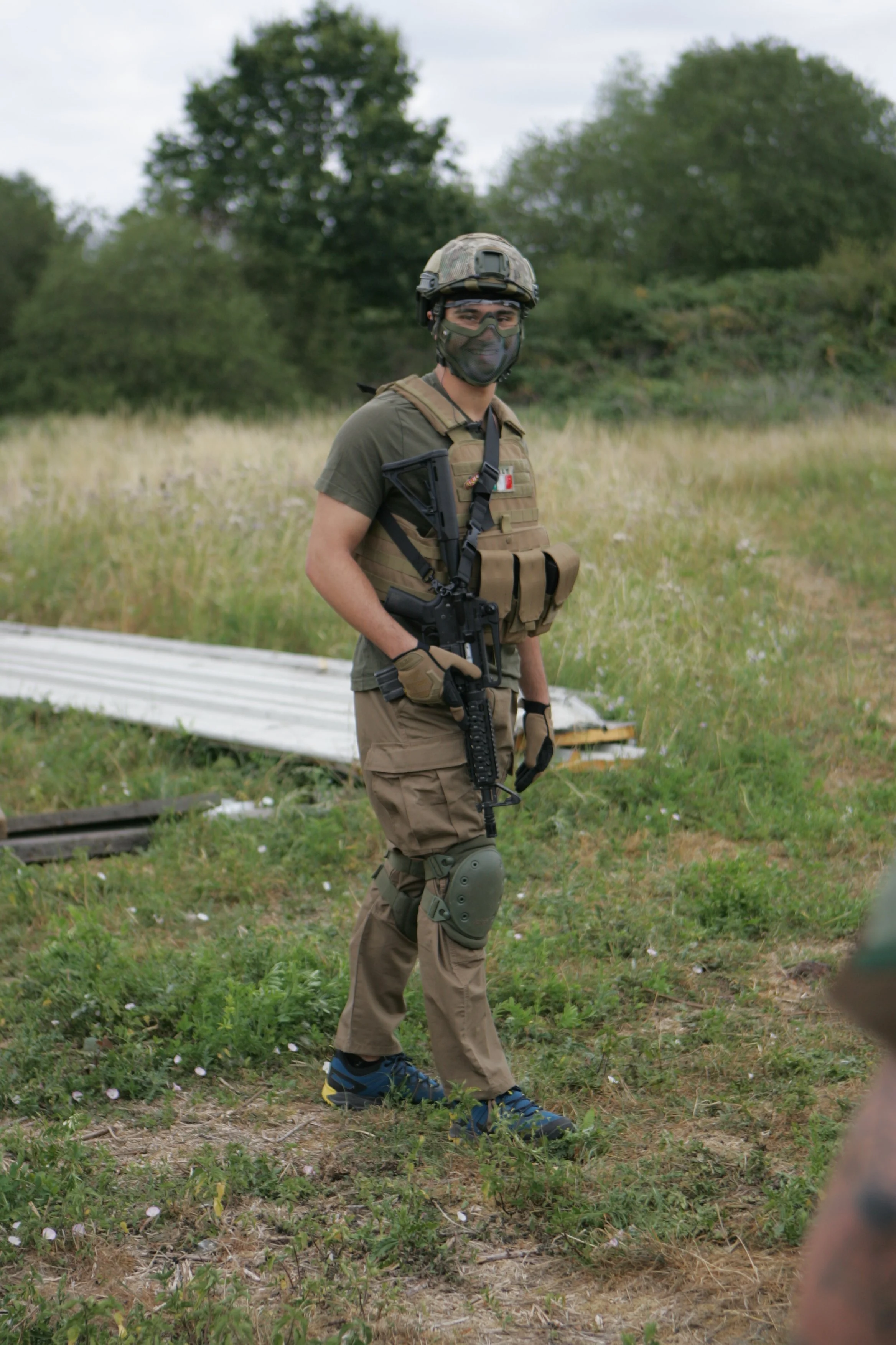 A man dressed in military tactical gear, including a helmet, gloves, and knee pads, stands outdoors in a grassy field with trees in the background. He is smiling and looking at the camera, holding a rifle.