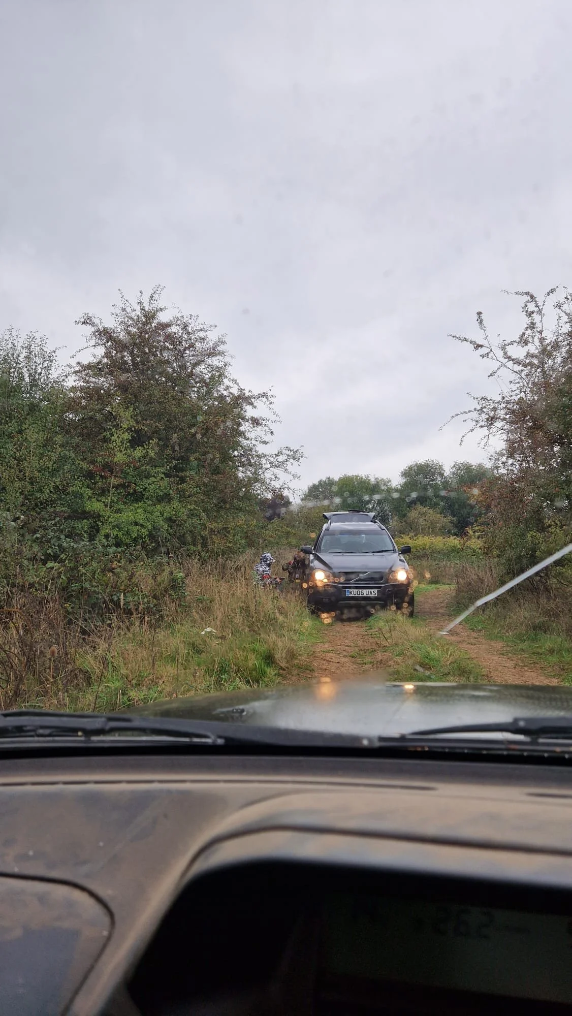 View from inside a car showing a muddy dirt road with grass and bushes on both sides. In the distance, a black SUV is stopped on the path, and a person appears to be near motorcycles.