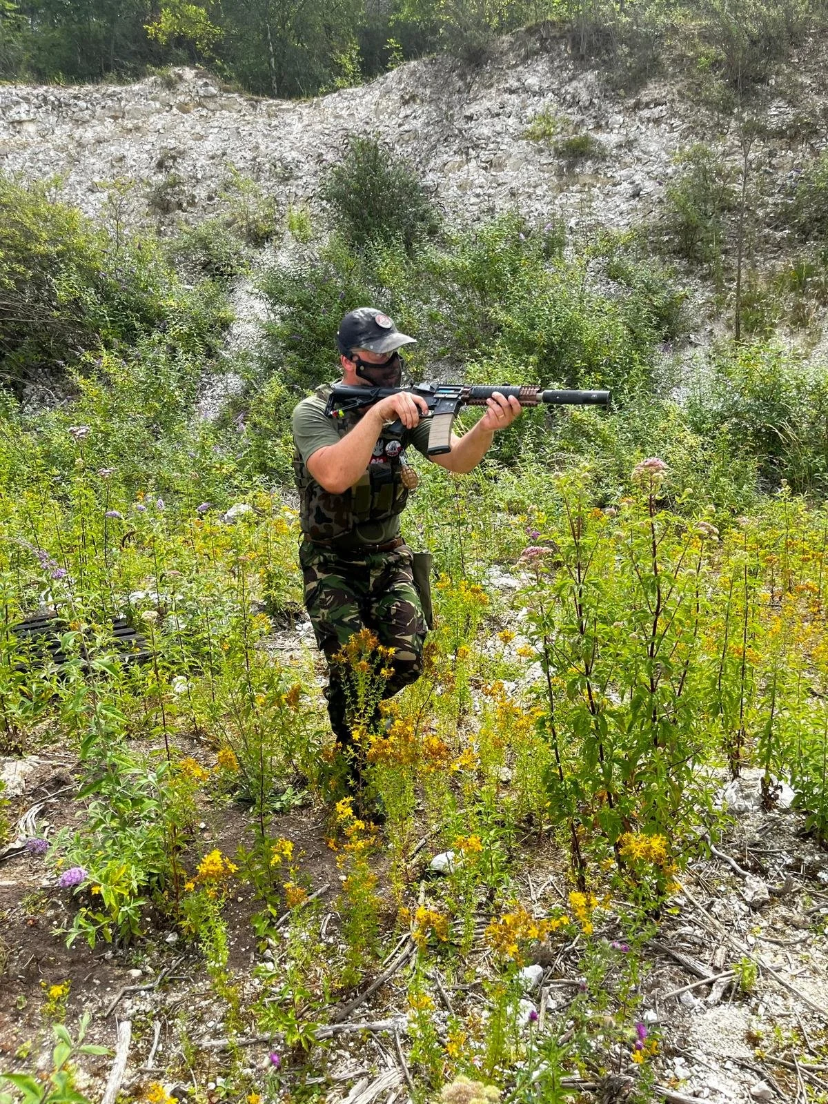 A person wearing camouflage pants, a green shirt, and a tactical vest, is aiming a rifle in a wooded outdoor area with tall plants and a rocky hill in the background.
