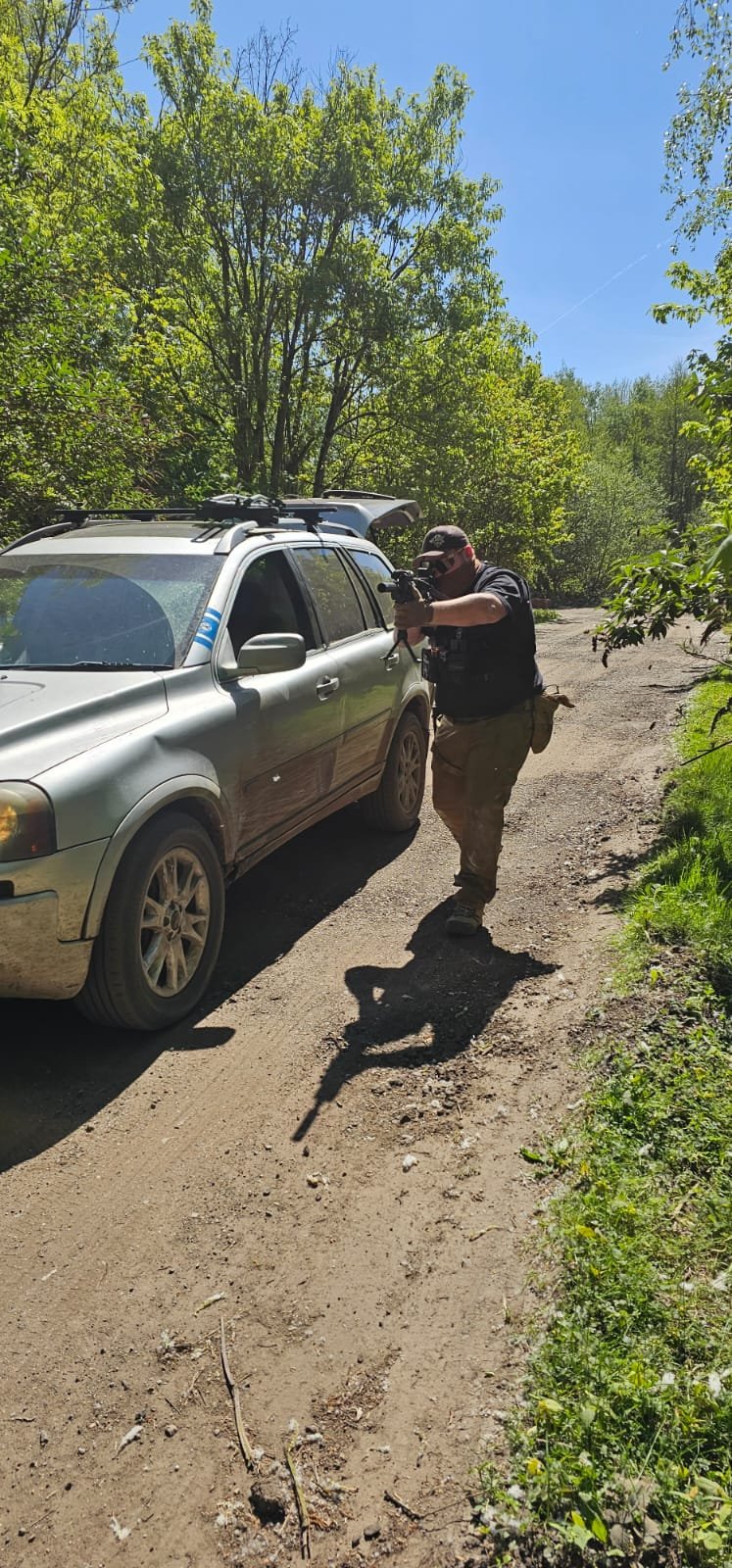 A man in tactical gear aiming a rifle while standing next to a gray SUV on a dirt road surrounded by trees and greenery on a sunny day.