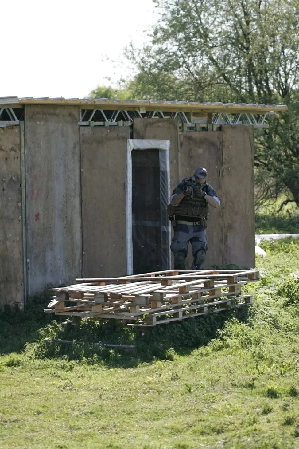 A tactical police officer in black gear and helmet standing outside a construction site with plywood walls and a pallet, holding a rifle.