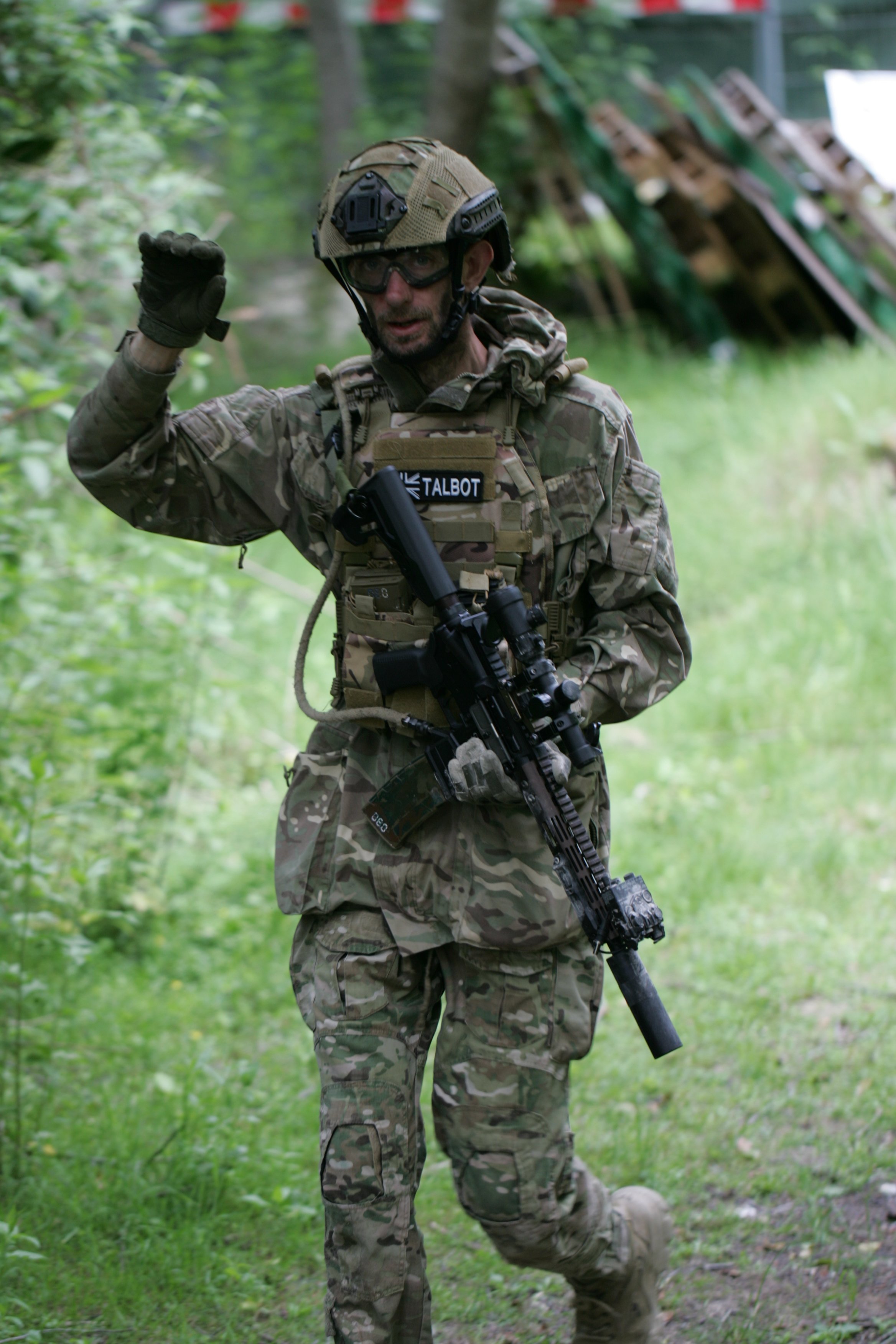 A soldier in camouflage uniform and helmet, with goggles, standing in a wooded area, holding a firearm, with greenery and trees in the background.