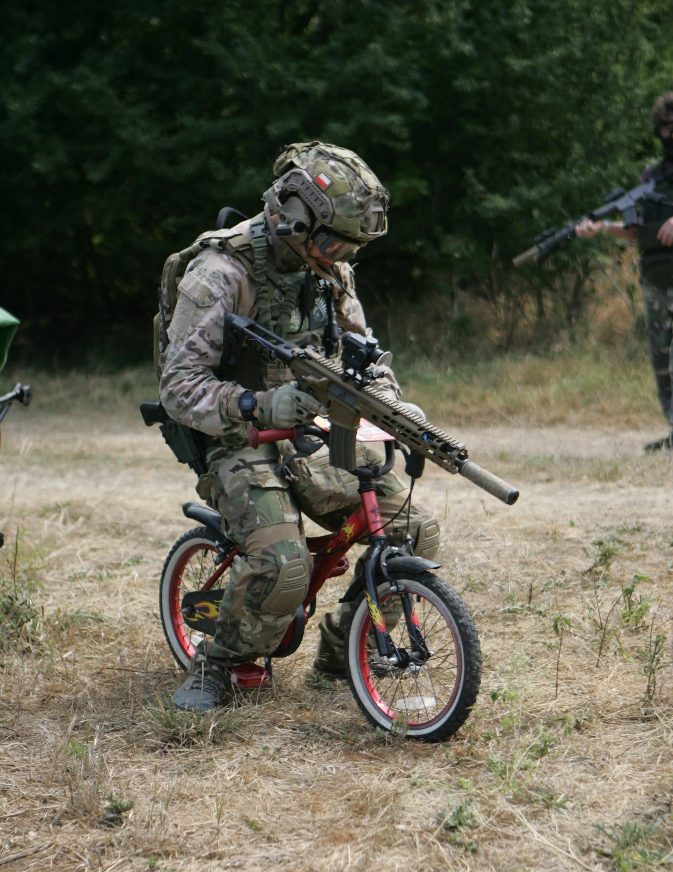 A soldier dressed in camouflage military uniform and helmet, kneeling with a rifle, involving a young child riding a small red bicycle with training wheels on a grassy, wooded area.